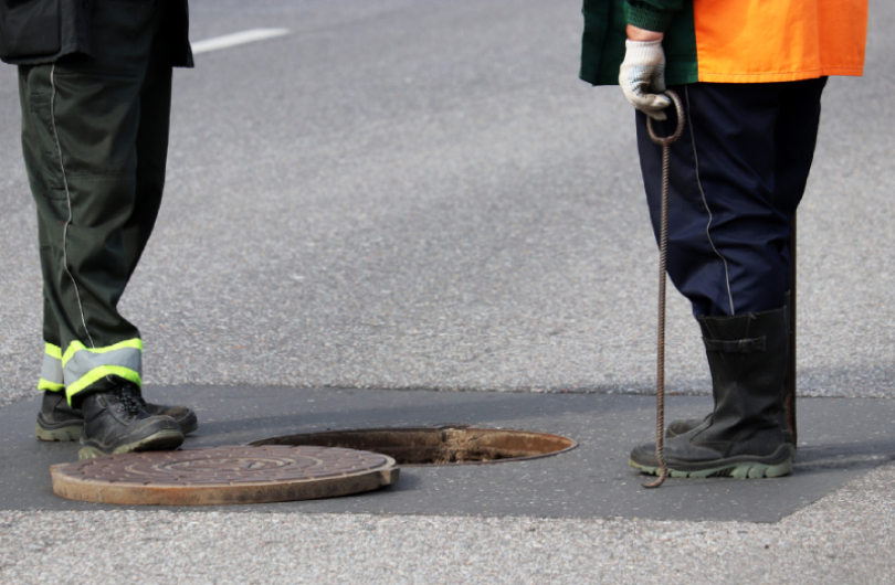 Two people are standing by an open sewer hatch in the asphalt. One of them holds a tool used to lift the heavy lid.