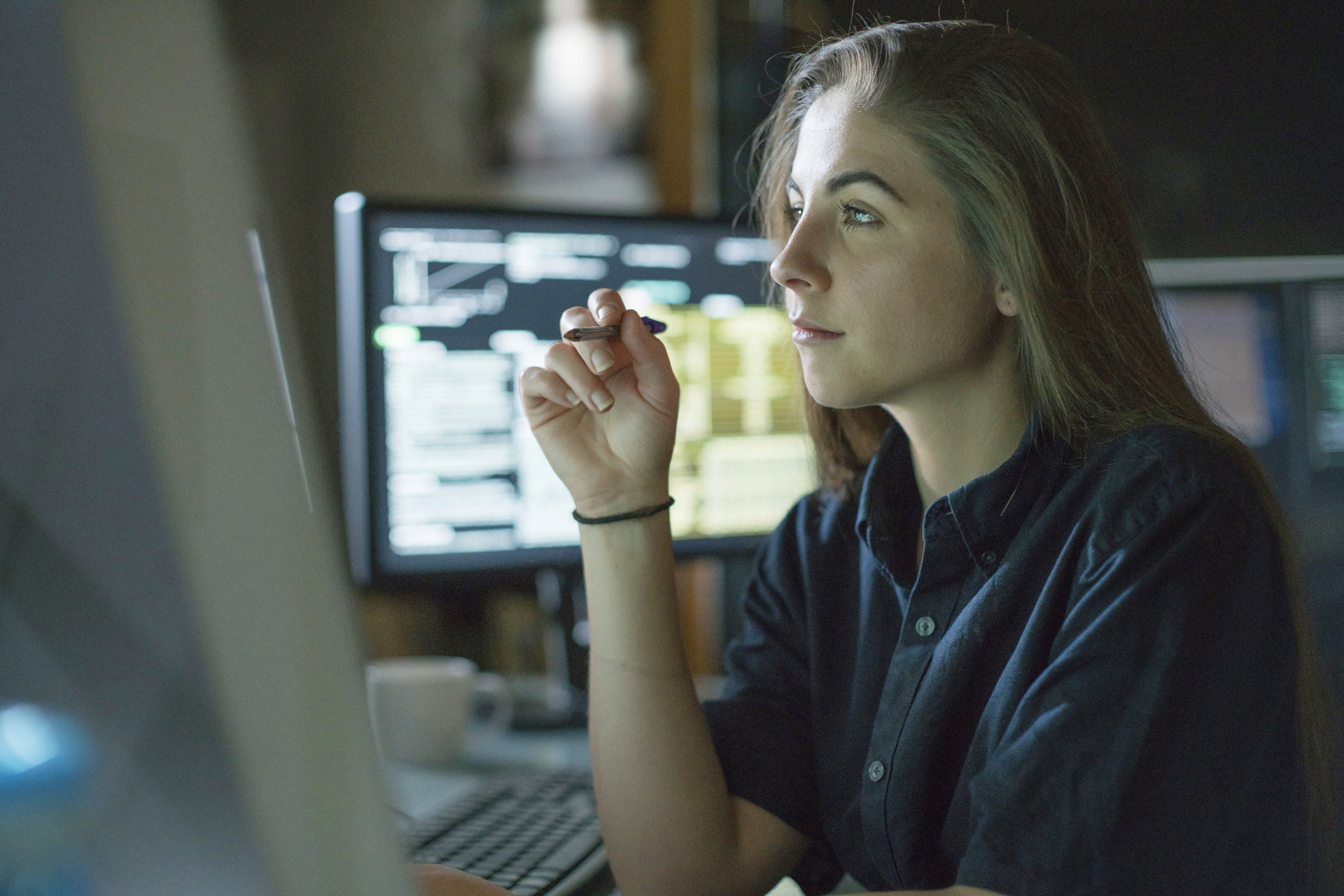 A woman with long hair, wearing a dark shirt, sits focused in front of a computer screen, holding a pen. Symbolizes concentrated work that can be linked to competence development and career advancement.