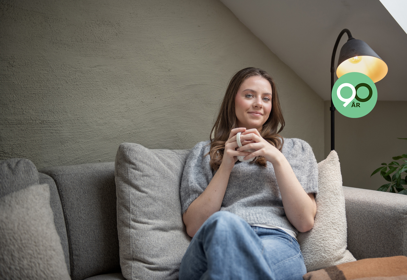 Ung kvinne sitter i en lys sofa og smiler med en kaffekopp i hendene