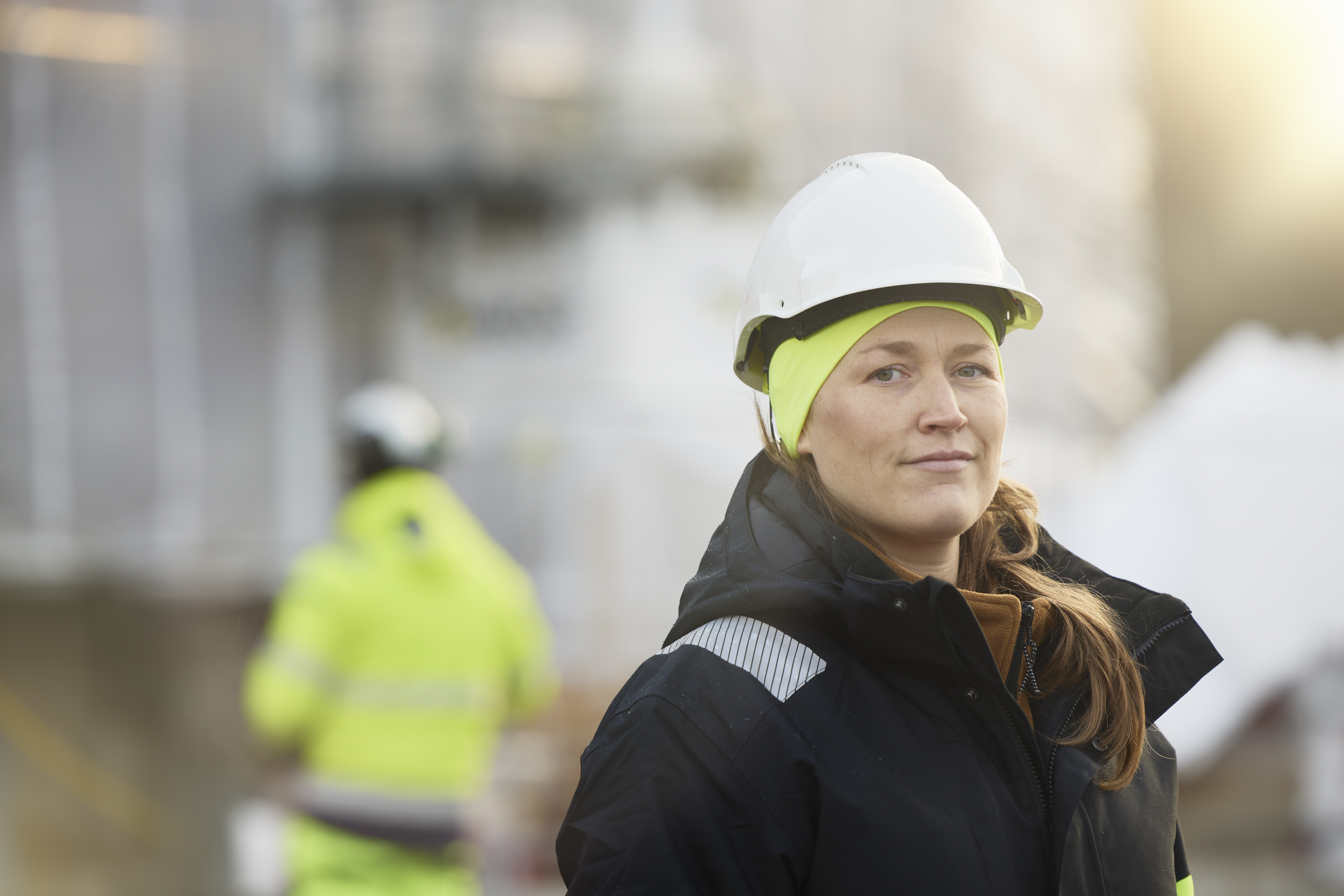 A smiling female industrial worker wearing protective equipment and a work jacket stands on a construction site.