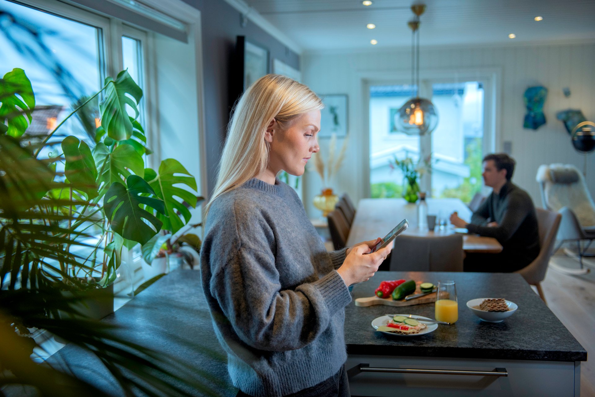 A woman who is anxiously reading on the phone at home, a man is sitting at the dining table in the background and looking out into the sky.