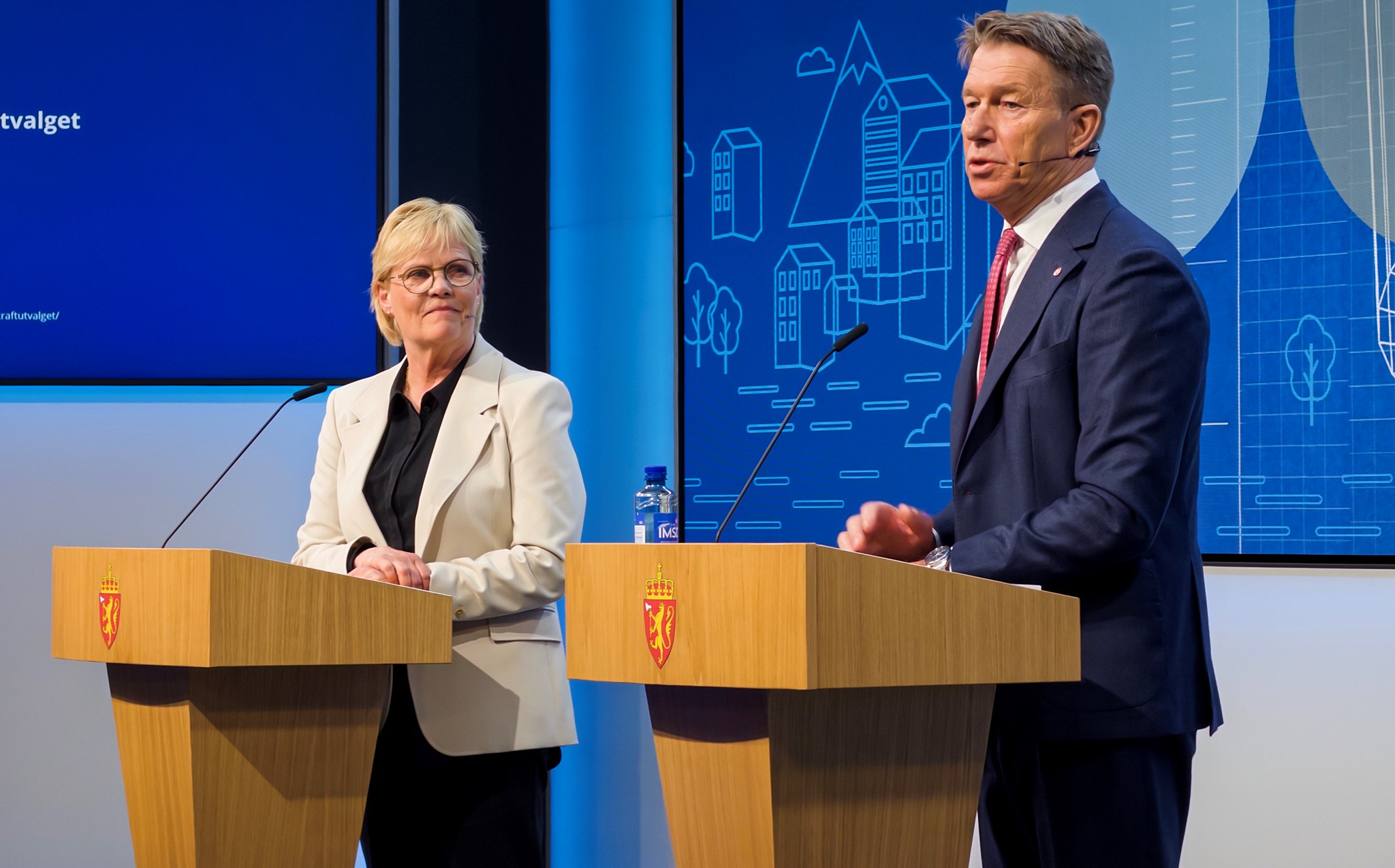 To personer står ved talerstoler med Norges riksvåpen. En skjerm med illustrasjoner av bygninger vises i bakgrunnen. Foto: Jo Henrik Jarstø/Energidepartementet 