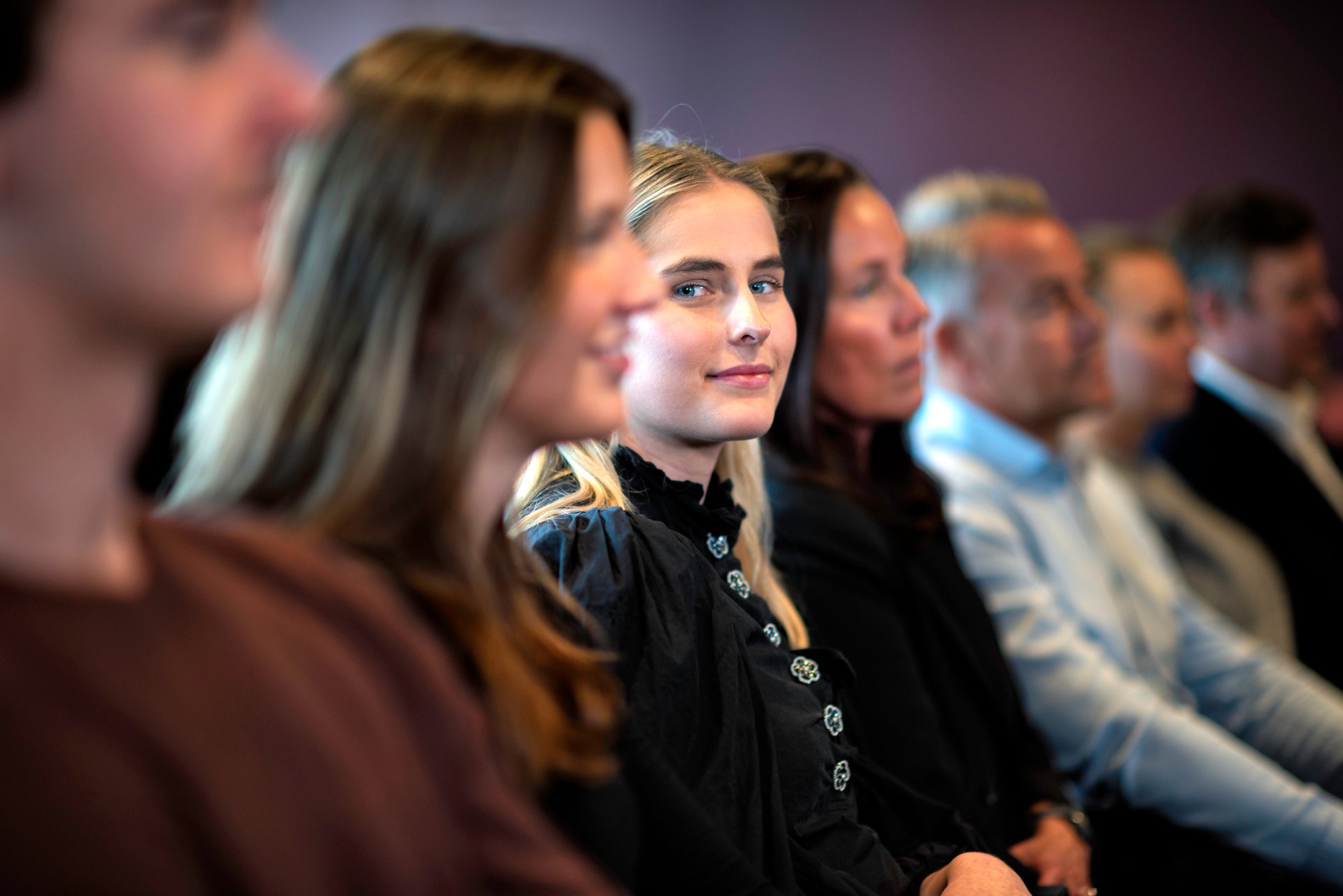 Ansatte sitter bortover på en rekke. Ung jente ser i kamera og er i fokus. 