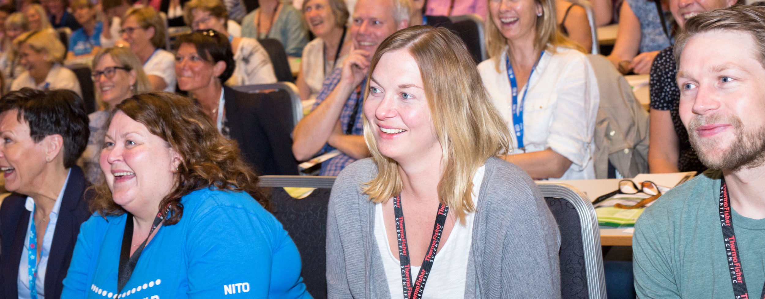 The picture shows smiling course participants sitting in a hall. The atmosphere is engaged and positive.