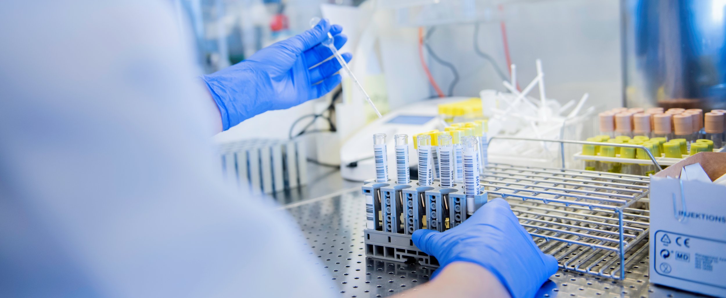 A person with blue gloves works in a laboratory, handling test tubes in a rack. The background shows laboratory equipment.