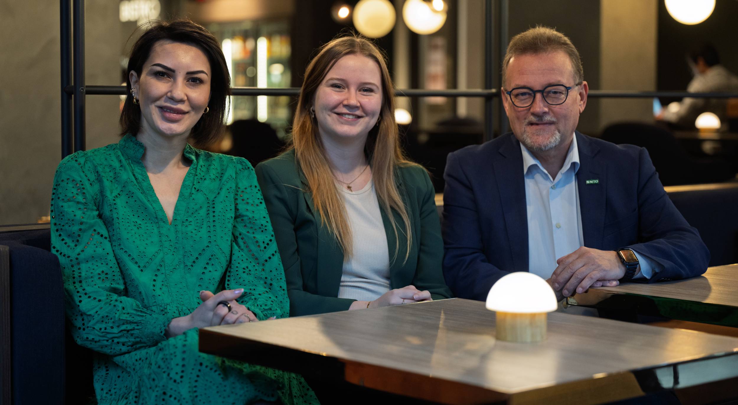 The photo shows three people sitting side by side in a modern meeting area with dim lighting.