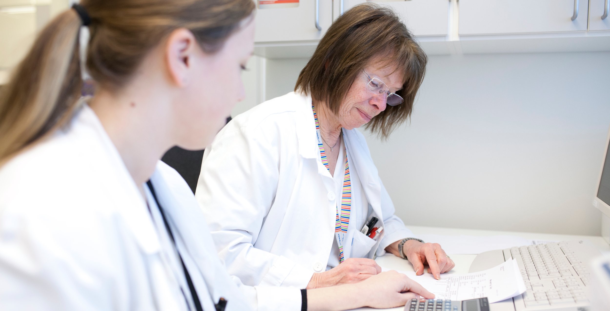 Two people in white coats work together at a desk with documents and a computer in a healthcare office environment.