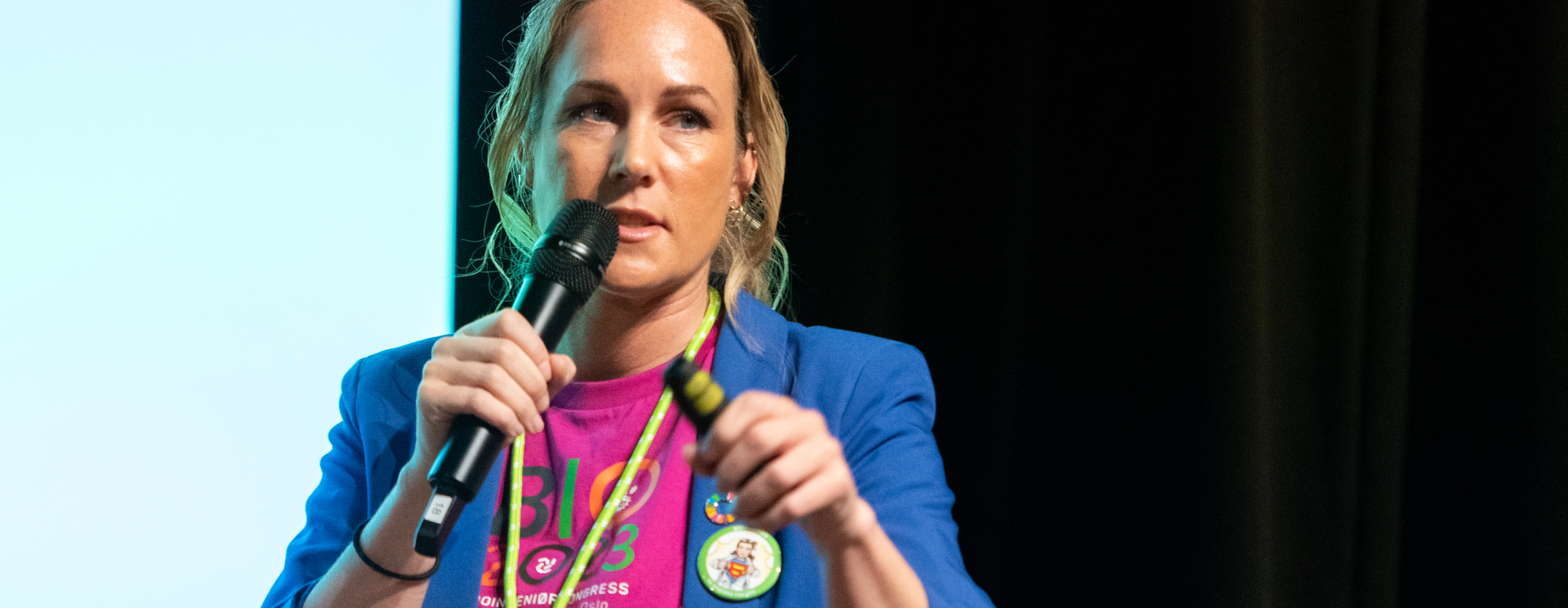 The image shows a woman holding a microphone, wearing a blue jacket, during a presentation or at a conference.