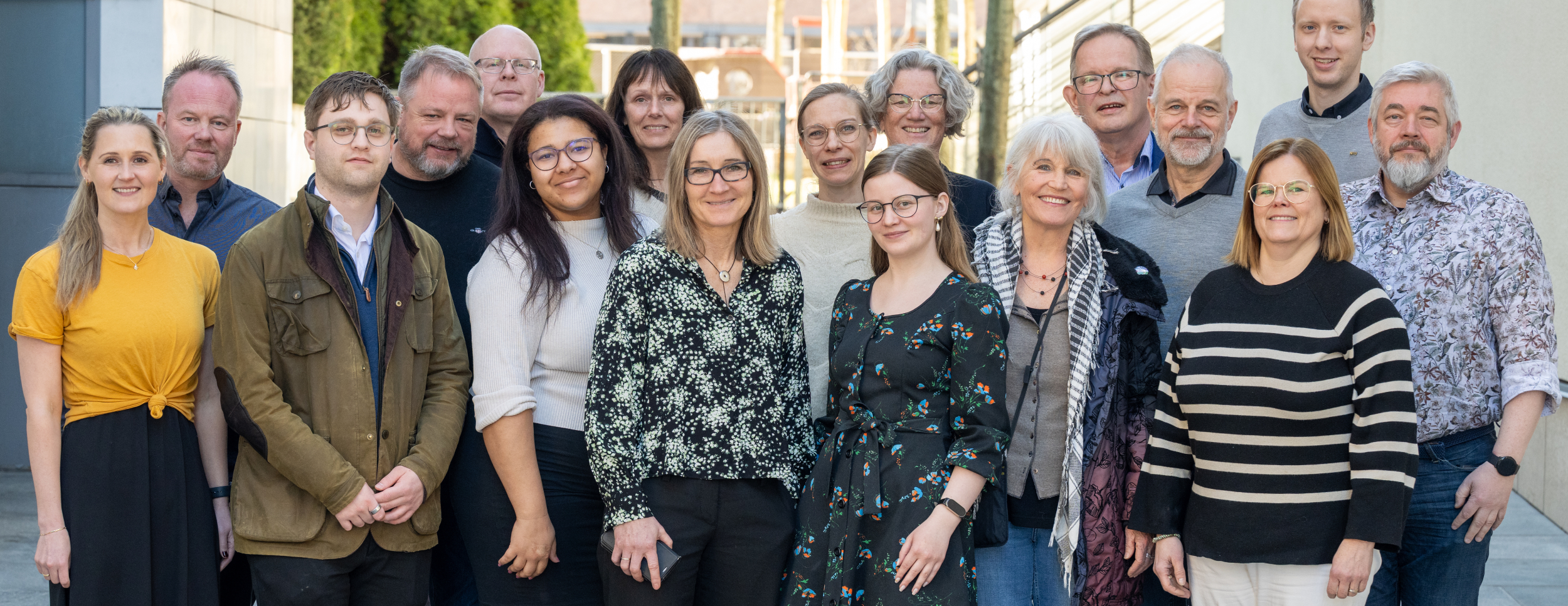 The picture shows 18 adult women and men standing outside in daylight and smiling at the camera. They are dressed casually, with buildings and trees in the background.