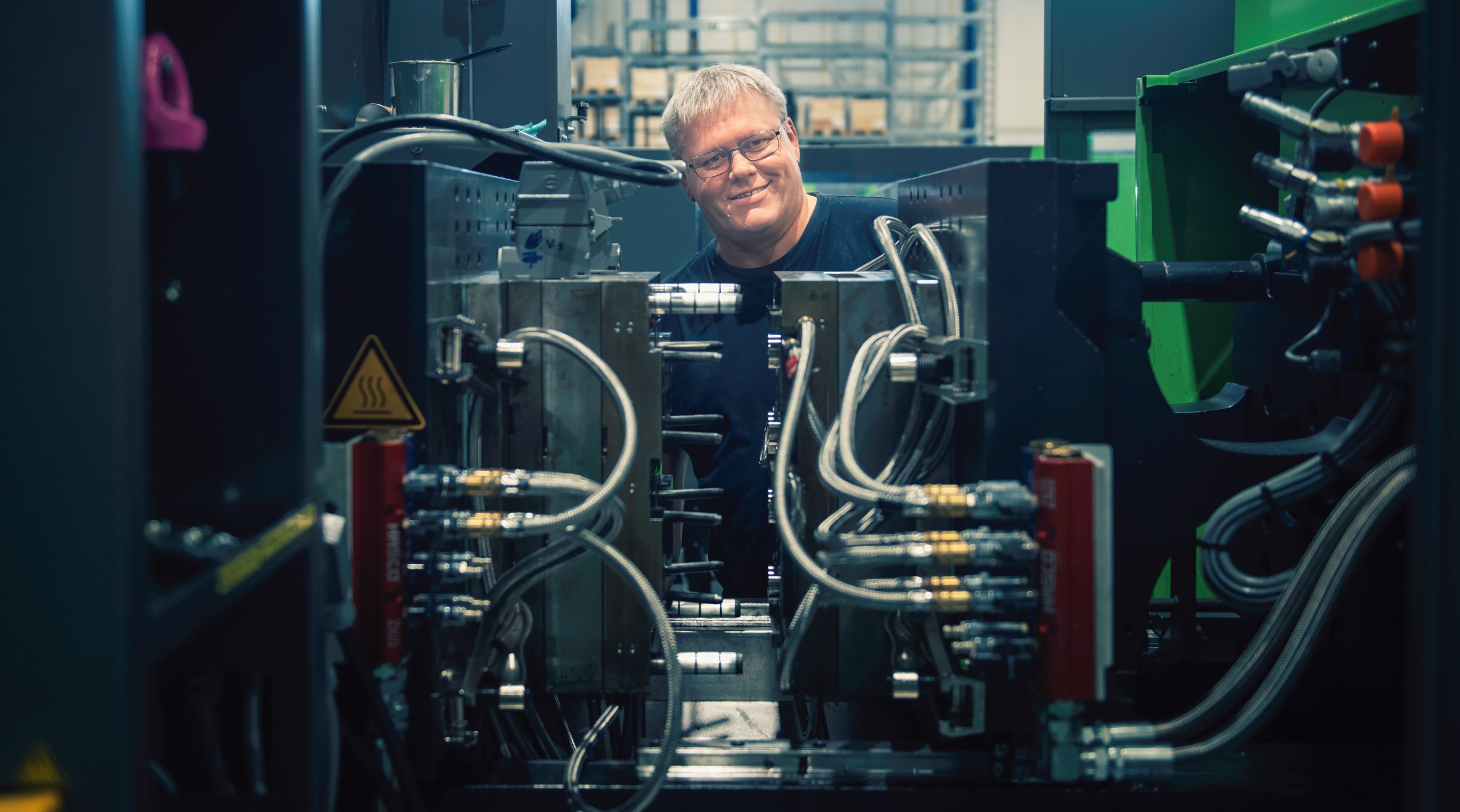 The picture shows a smiling man with glasses standing behind a large industrial machine, surrounded by metal pipes and hydraulic hoses.