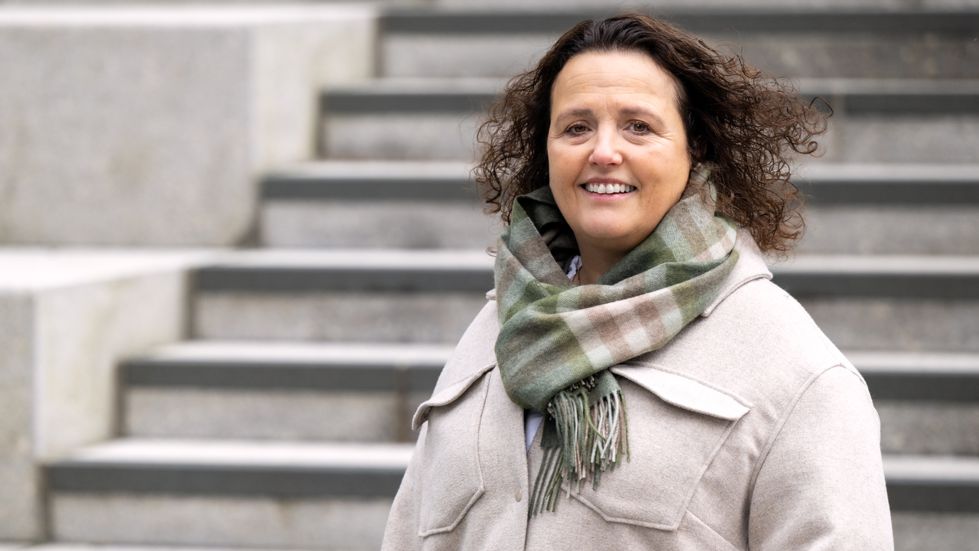 En kvinne med krøllete hår, lys frakk og rutete skjerf står foran grå trappetrinn.