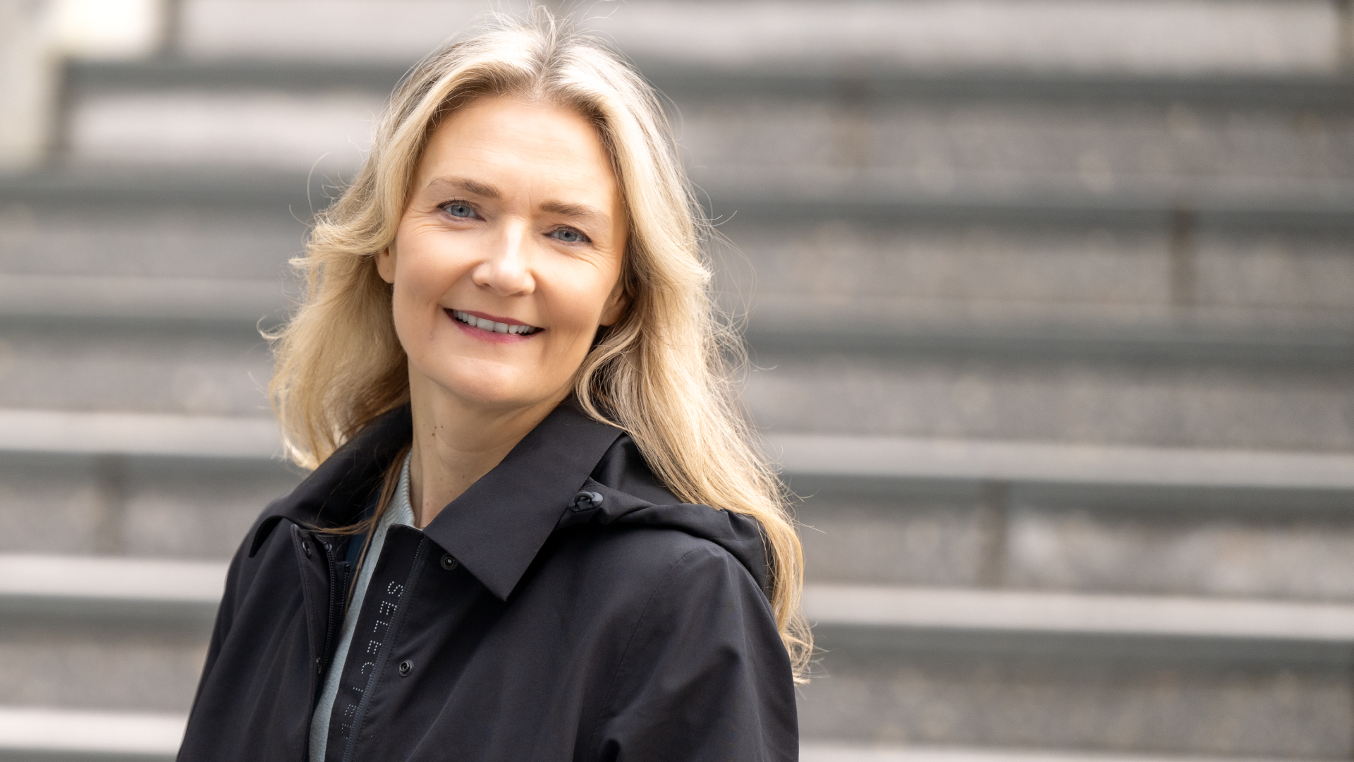 En kvinne med lyst hår iført mørk jakke står foran grå trappetrinn.