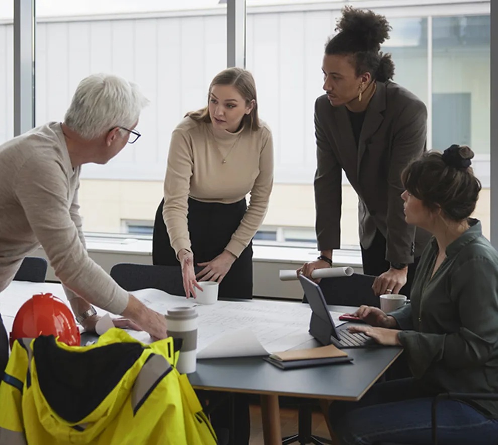 Fire personer står og diskuterer rundt et bord