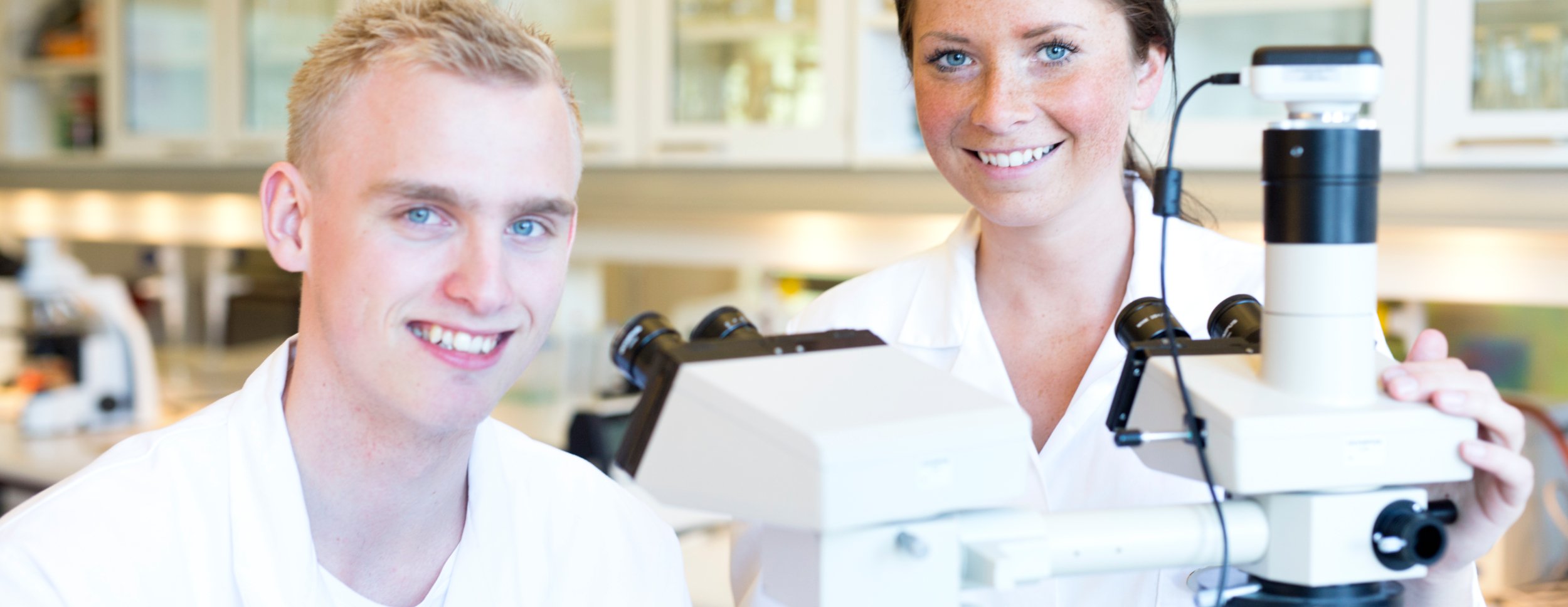 The picture shows two young biomedical laboratory technician students in laboratory coats under a microscope in a laboratory environment.
