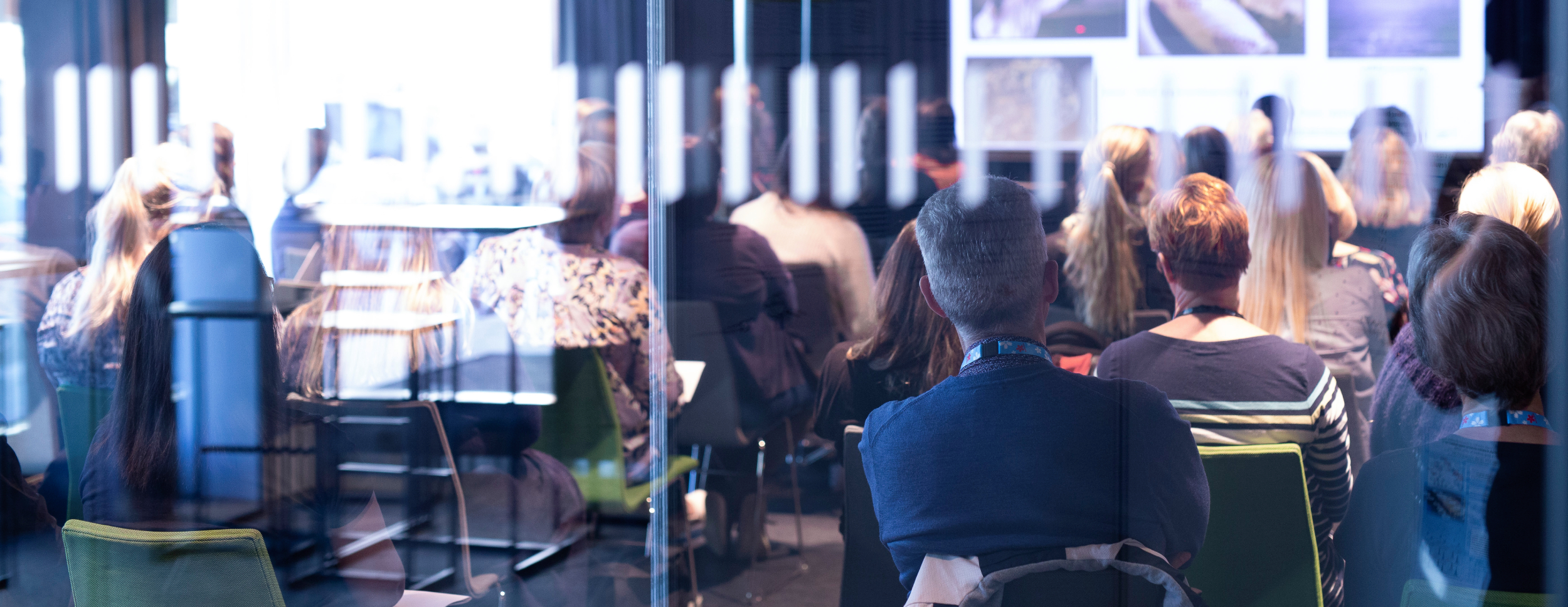 The audience watching a presentation in a bright conference room. The picture was taken from behind with blurry reflections in the foreground.