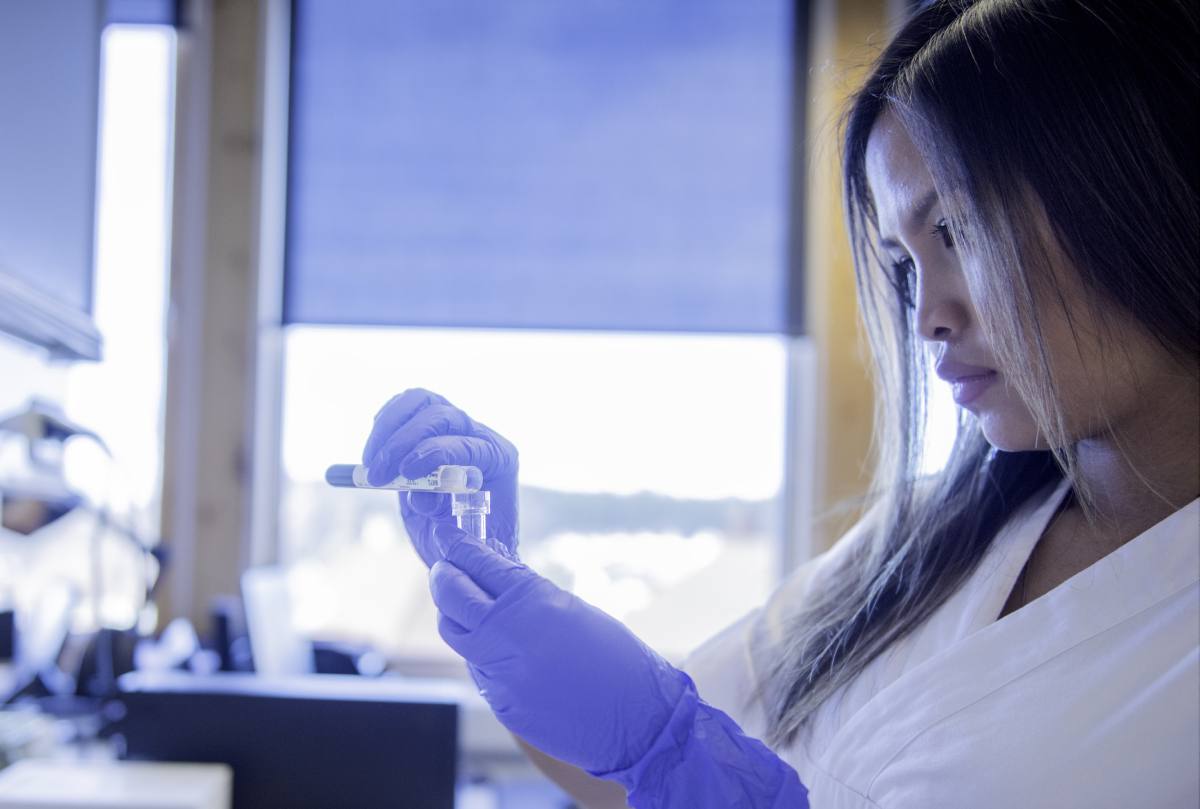 The photo shows a woman in a lab coat and blue gloves handling a small plastic container of liquid. She is in a bright laboratory.