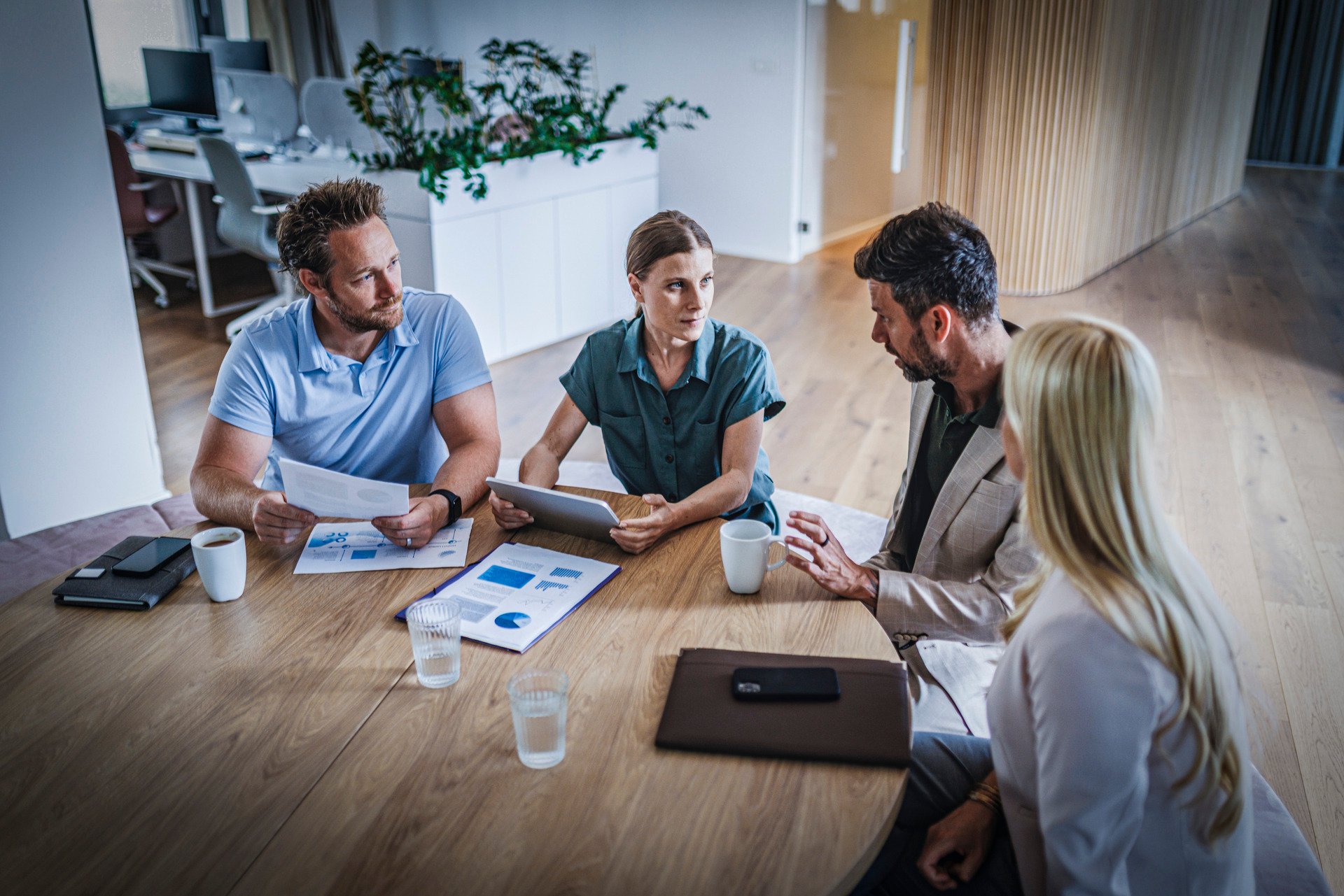 Four people have a meeting around a table in a modern office, with documents, laptop and coffee cups in front of them.