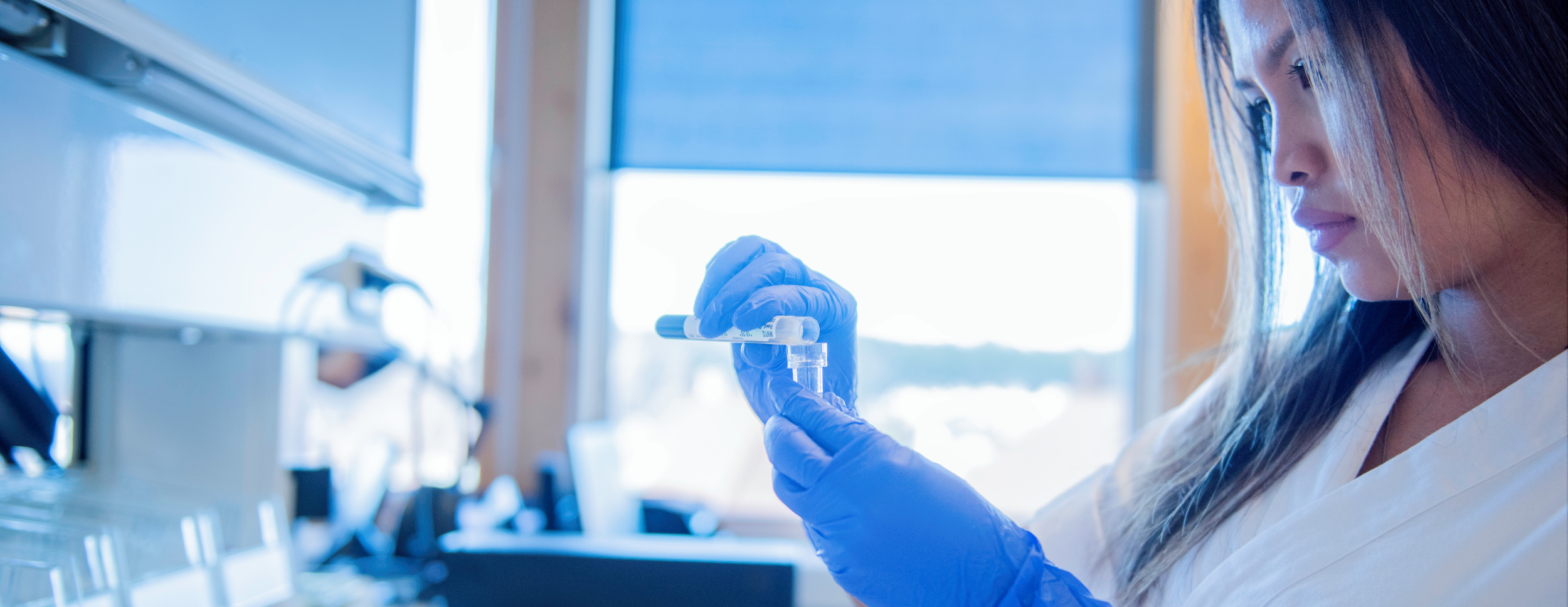 The photo shows a woman in a lab coat and blue gloves handling a small plastic container of liquid. She is in a bright laboratory.
