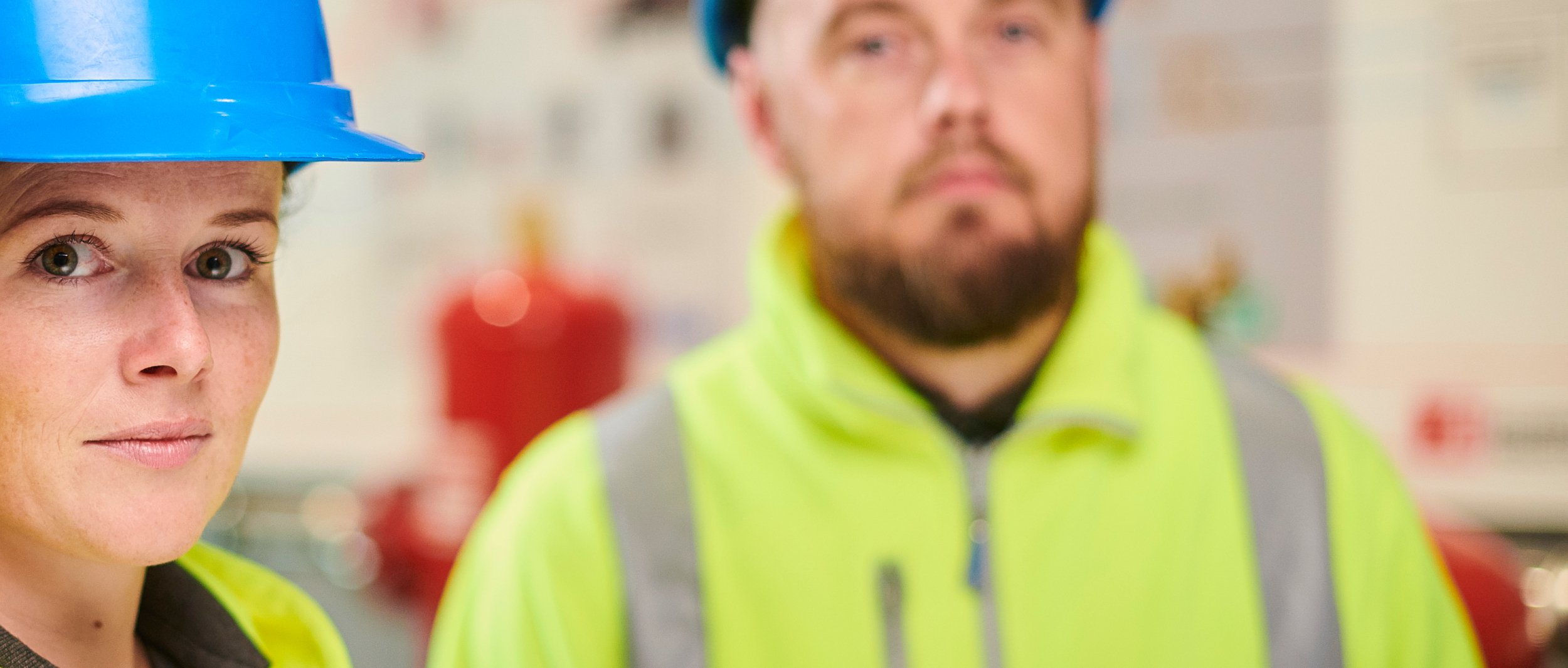 The picture shows two people in yellow work jackets with reflective stripes and a blue helmet, in a working environment with a blurred background.