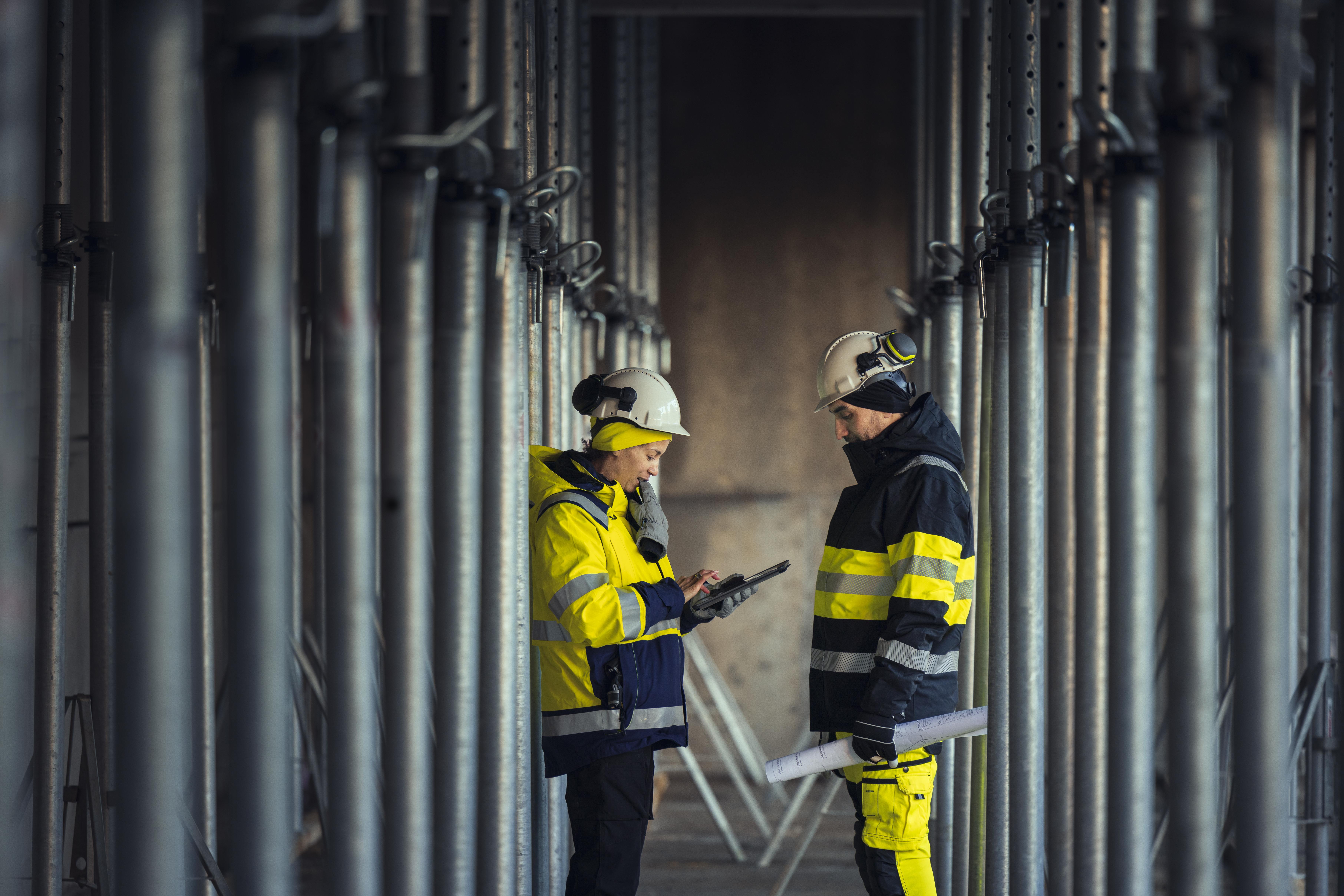  To ingeniører, mann og kvinne, iført arbeidstøy og hjelm snakker sammen i et anlegg med rør i bakgrunnen foto:GettyImages