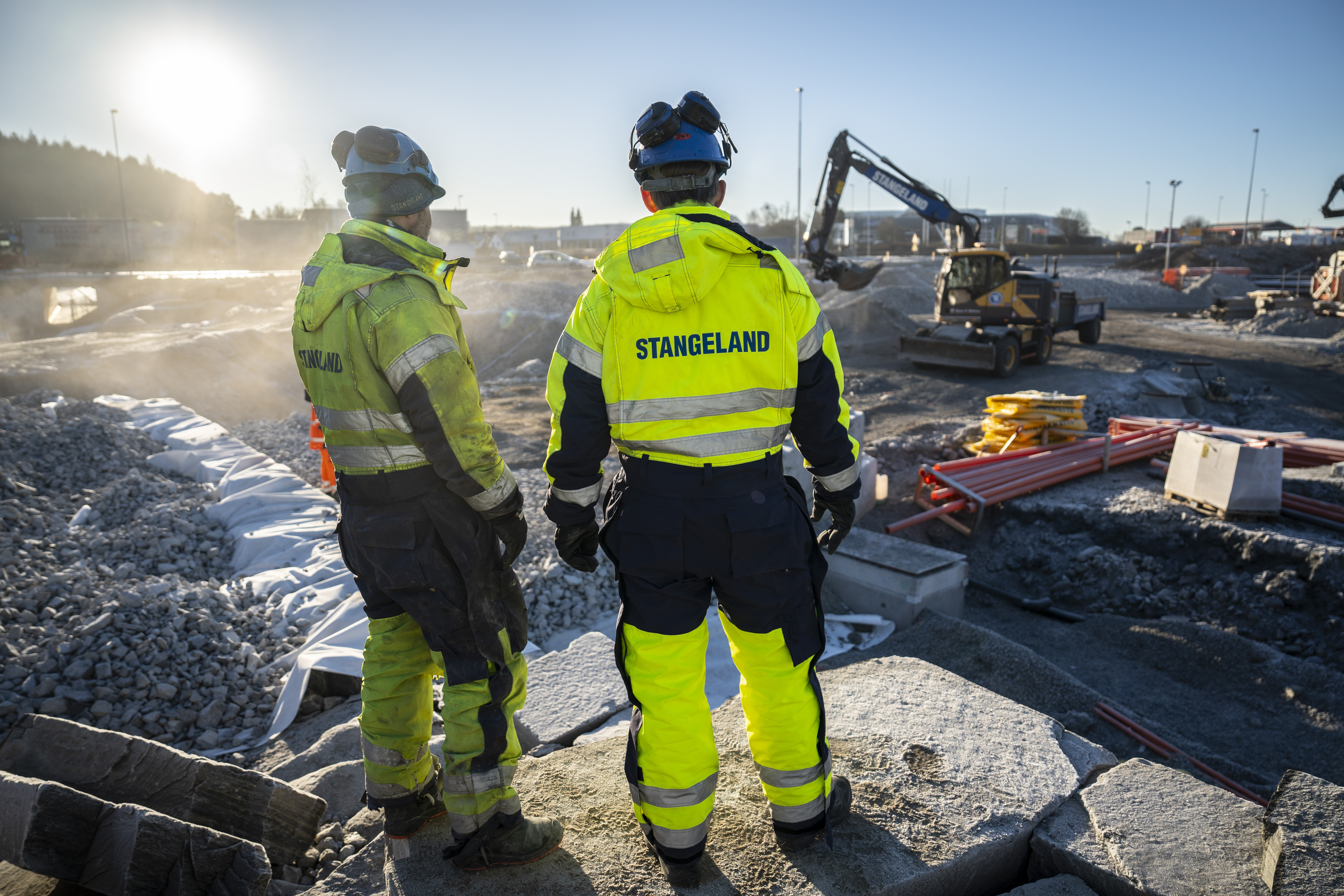 Bilde av to anleggsarbeidere som ser ut på et veiprosjekt. Gravemaskin og bygningsarbeid i bakgrunnen.