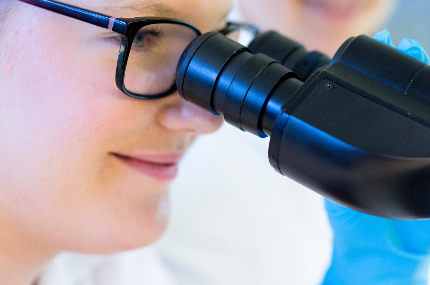 A person with glasses looks intently into a microscope. The image shows details of the face and the microscope.