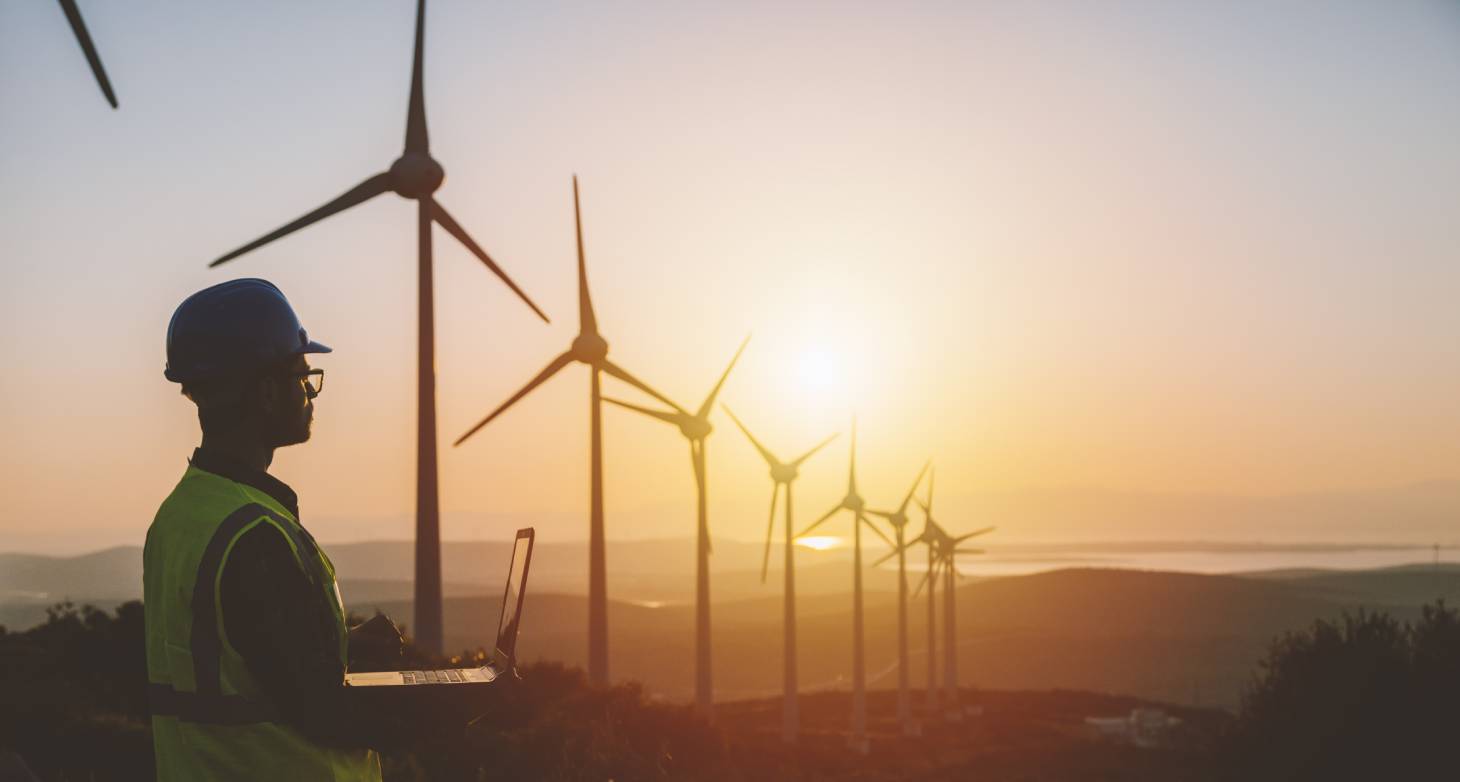 Siluett av ung mannlig, ingeniør med  bærbar datamaskin som arbeider stående ved siden av vindmøller.