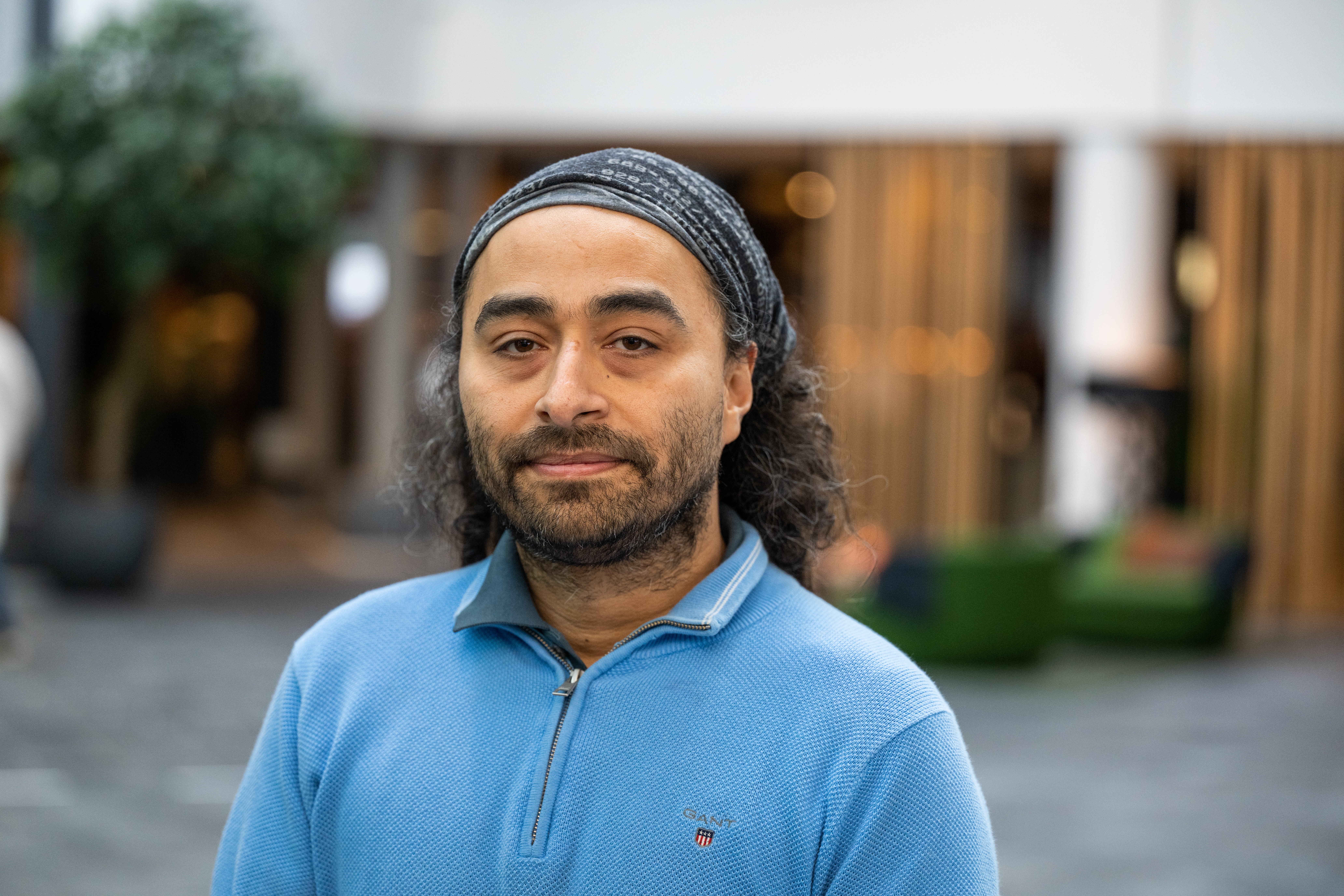 Portrett av en mann med langt hår og hodeplagg som står utendørs i et bymiljø.