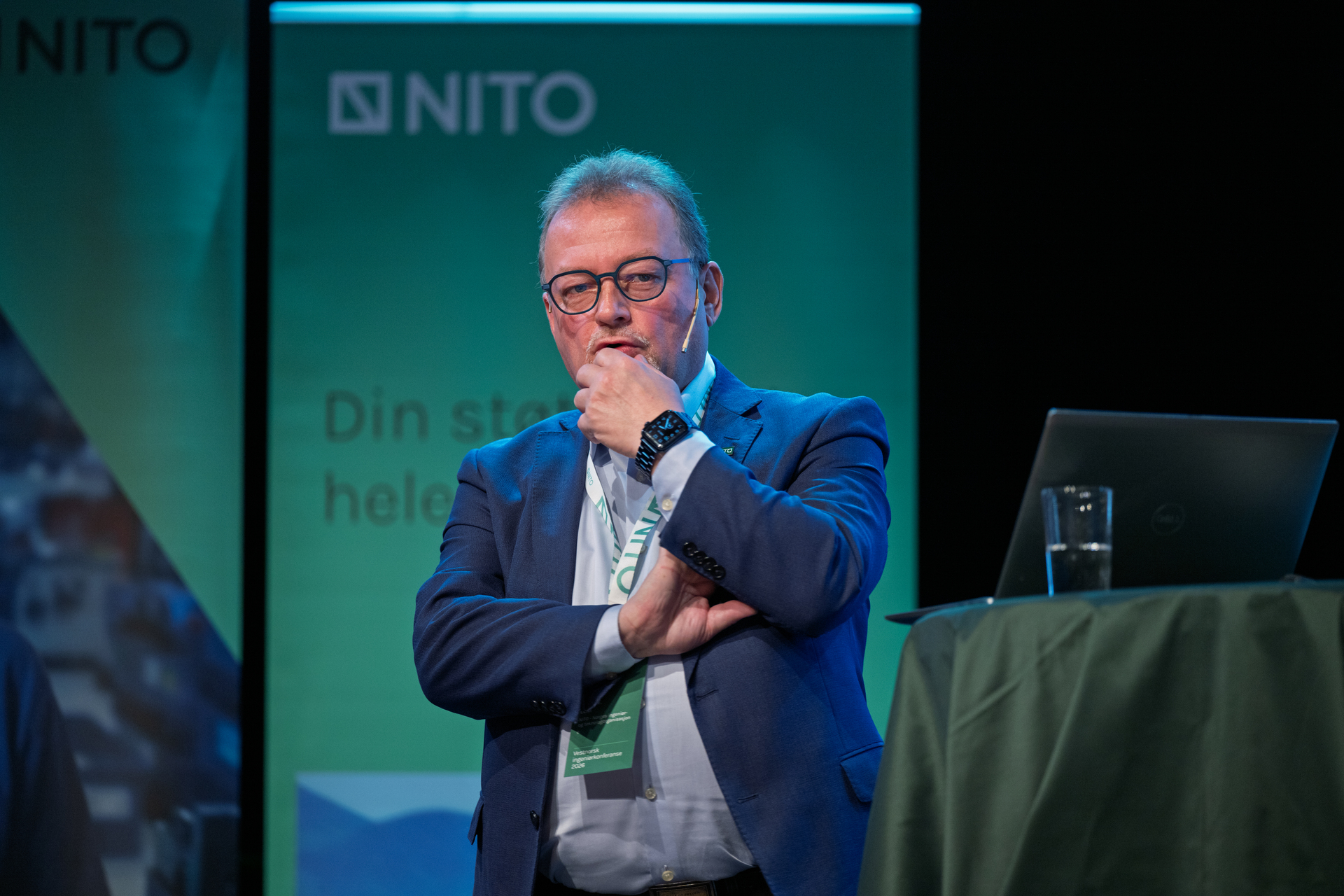Mann med briller iført skjorte og dress står på en scene