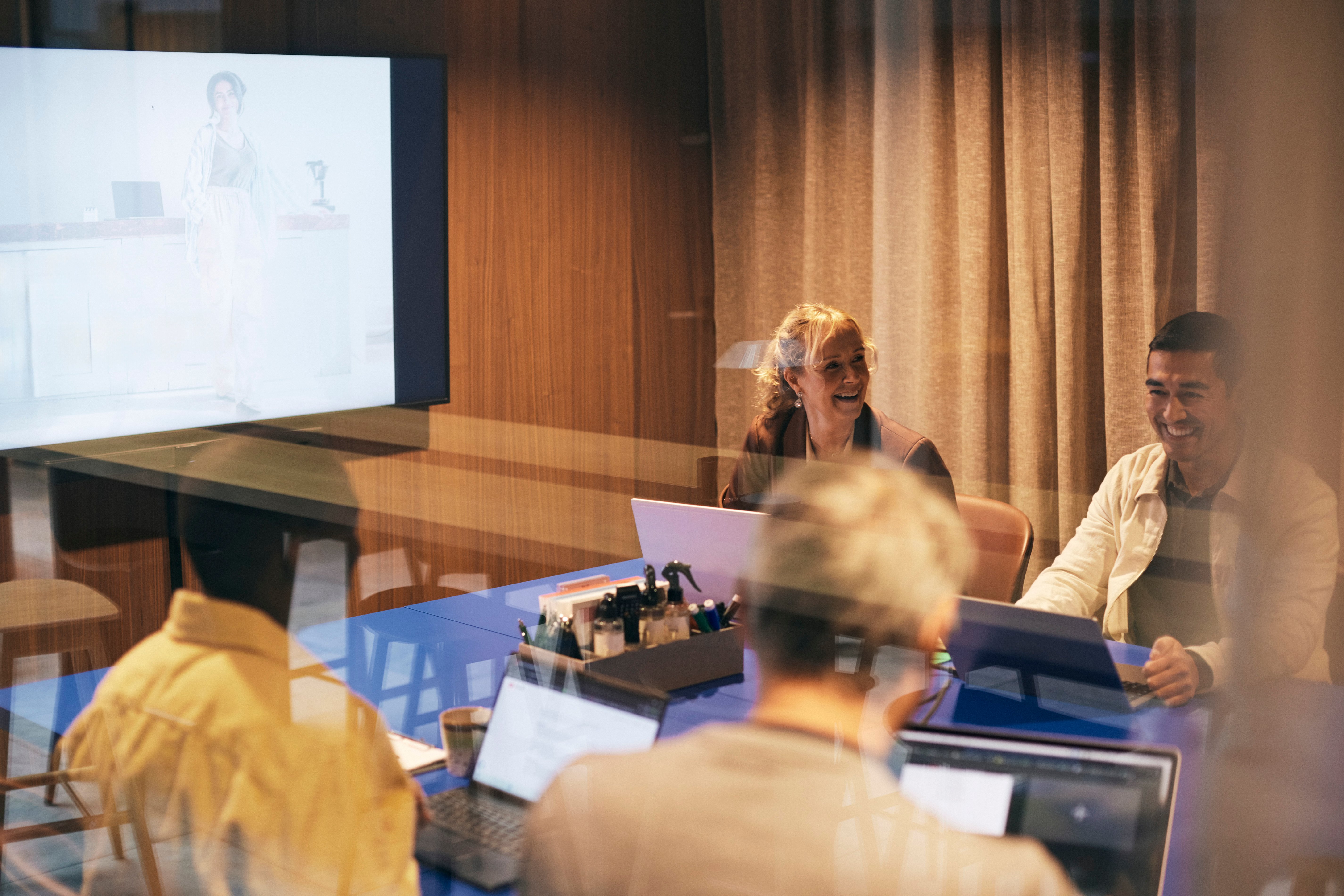 International collaboration meeting with participants around a conference table. One person presents on the big screen while participants follow along on laptops in a modern meeting room with wooden wall paneling. Photo: GettyImages