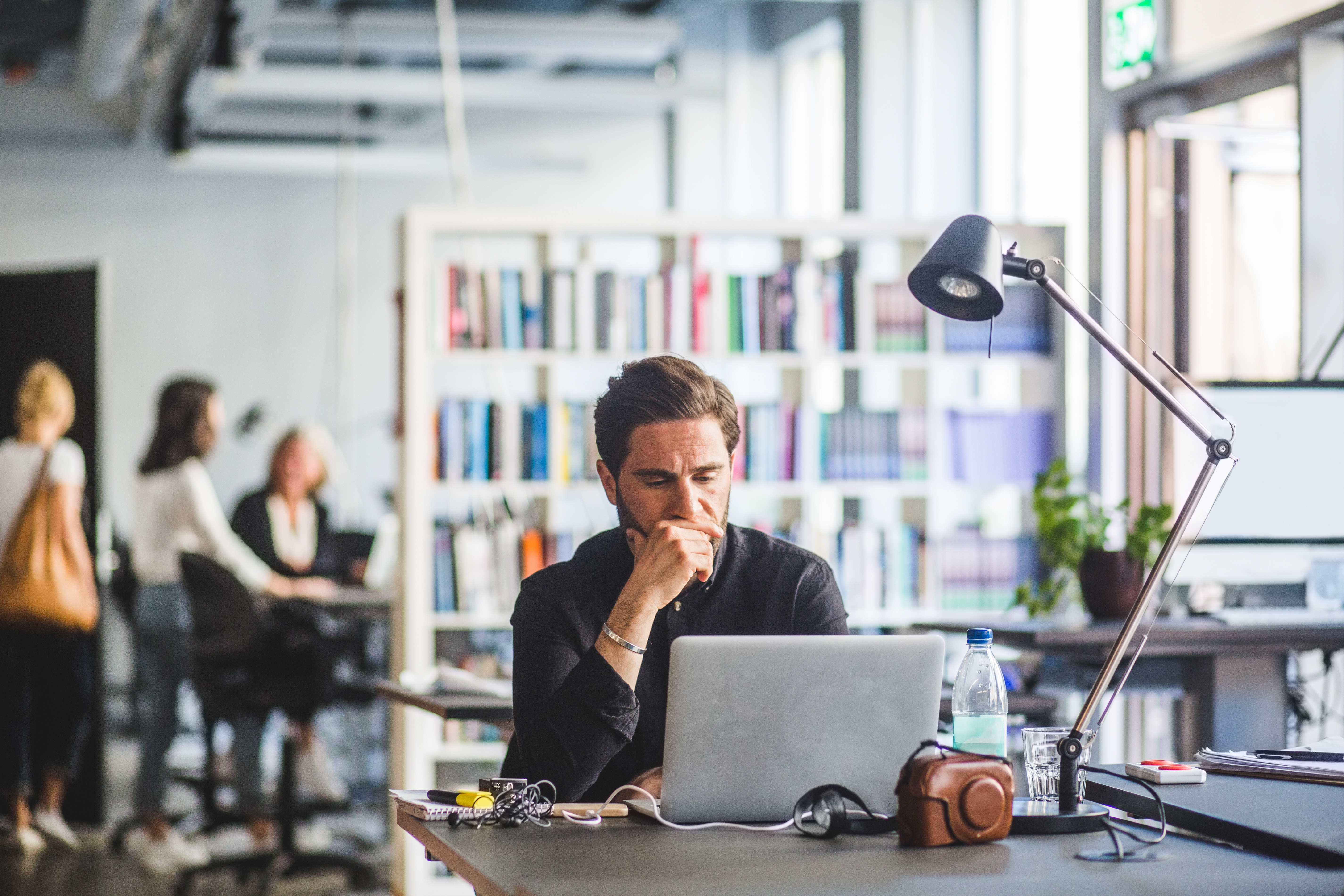 A man sits concentrated in front of a laptop in an office, with his hand on his chin, and seems to be reflecting or solving a problem. In the background, there are blurred colleagues and a bookshelf, indicating a dynamic work environment.