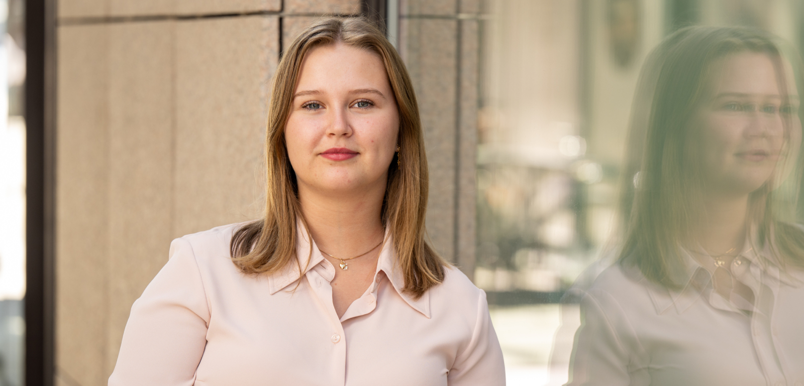 Picture of young woman with long brown hair and light pink blouse. The picture was taken outdoors.
