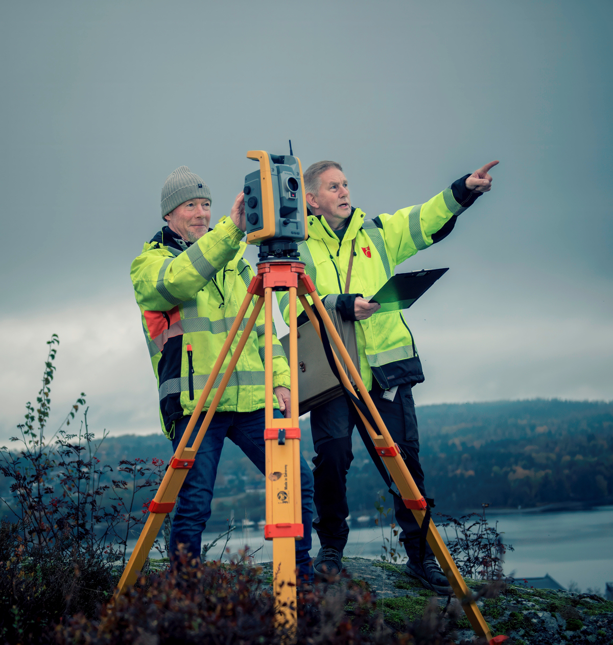 Two surveyors stand on a small hill with a theodolite and measure. In the background there are mountains and sea.