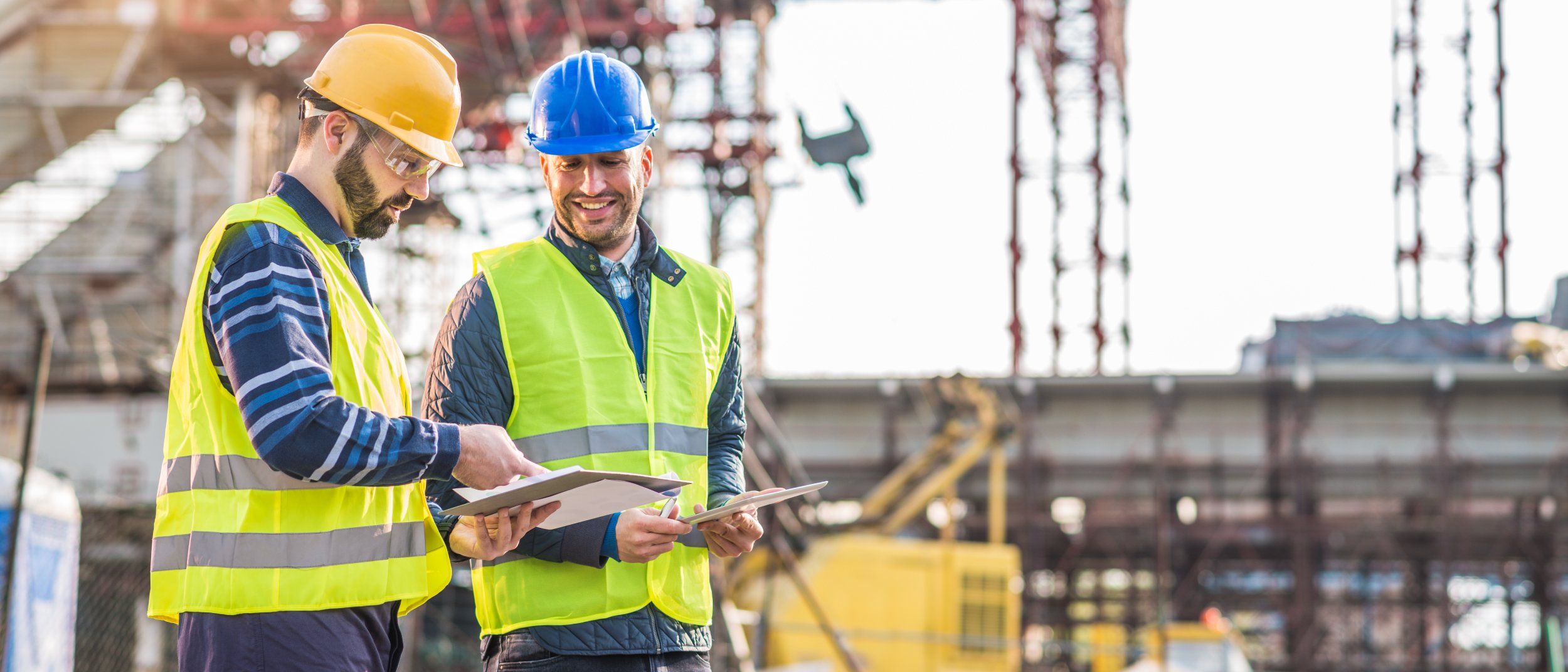 Two construction workers with yellow vests standing in the foreground of a large construction site with cranes in the background
