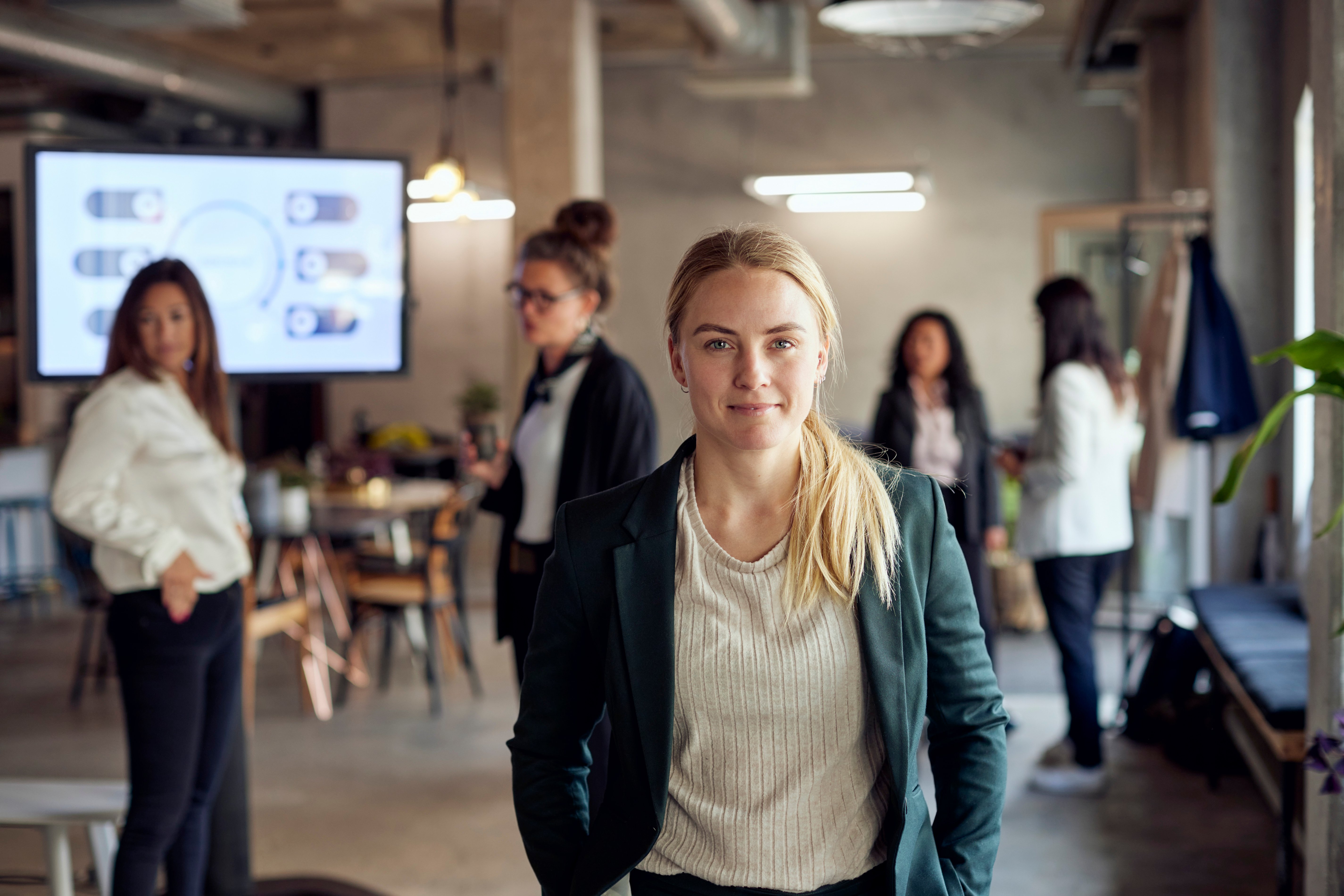 Confident woman in green blazer smiles in modern office environment with other colleagues in the background.