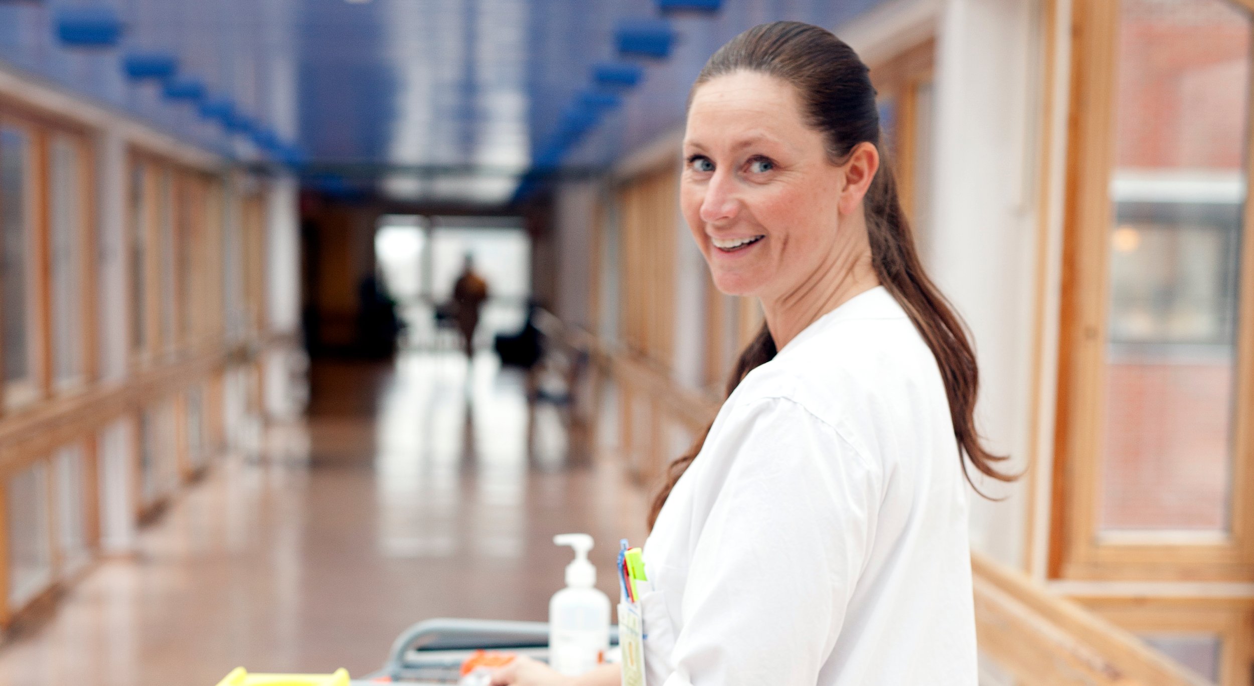 A woman in a white uniform stands in a bright hospital corridor with windows along the sides.
