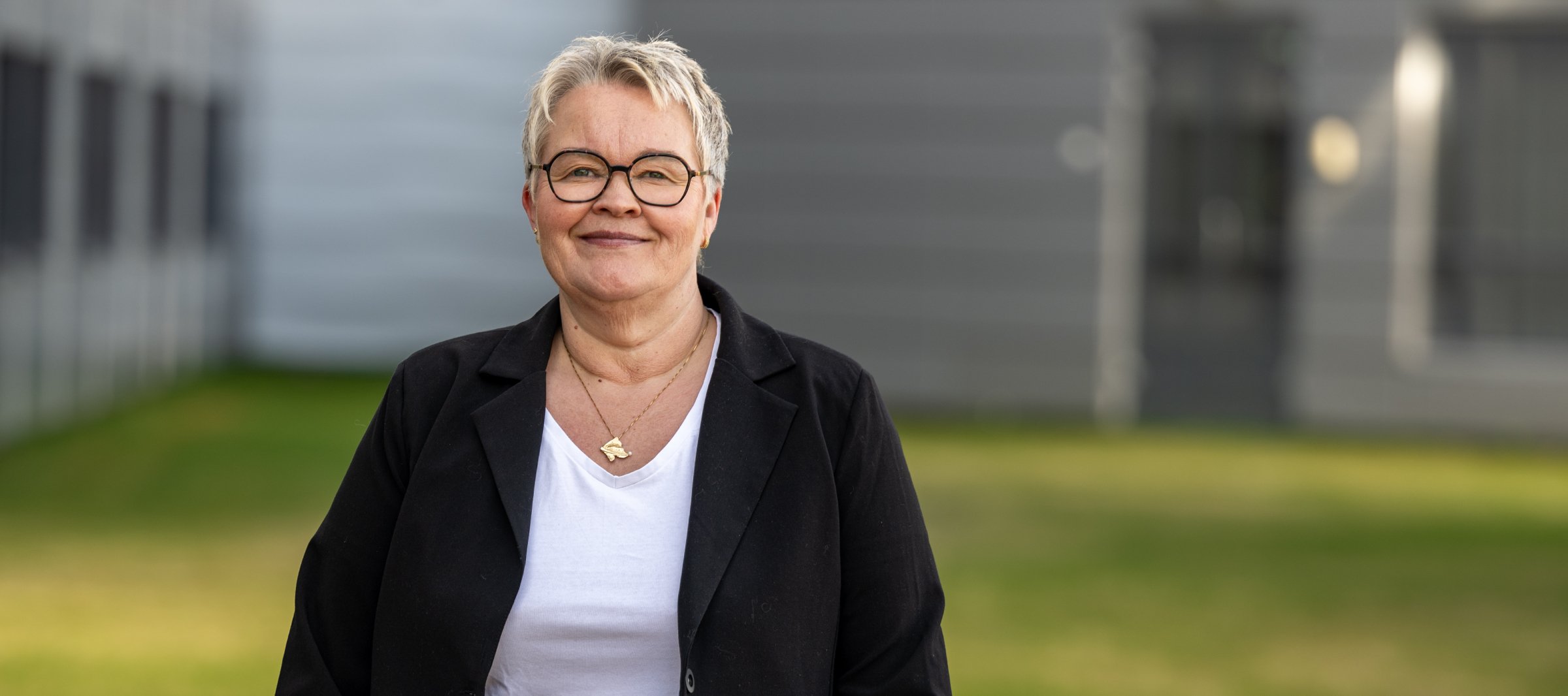 En smilende kvinne med kort, lyst hår, briller og svart jakke står utendørs foran en bygning med grønt gress i bakgrunnen.