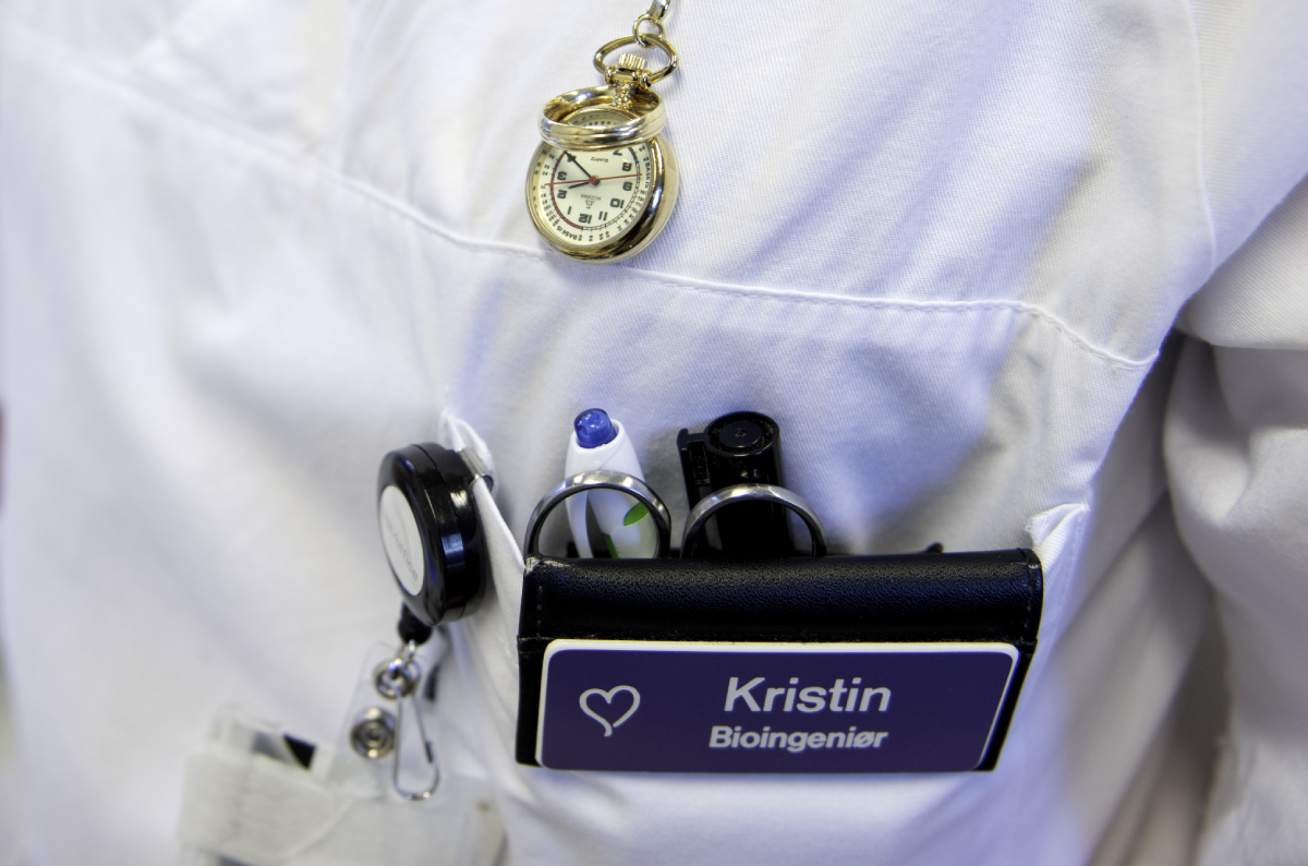 The photo shows the torso of a person in a white uniform. In the breast pocket there are pens, ID cards and a name tag with the text: "Kristin, Bioengineer".