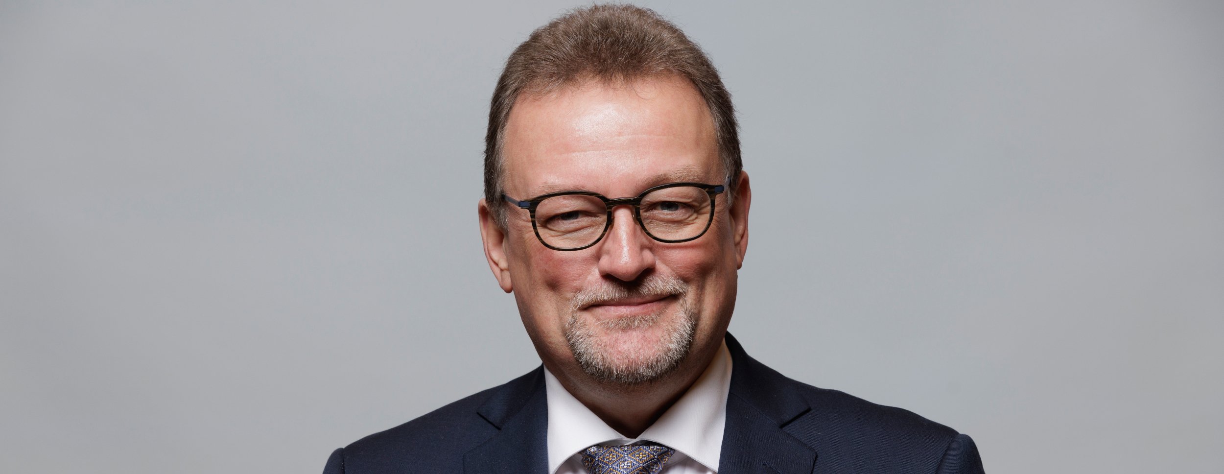 A smiling man in a suit and glasses looks straight into the camera. He has short, brown hair and stubble, and exudes security and friendliness.