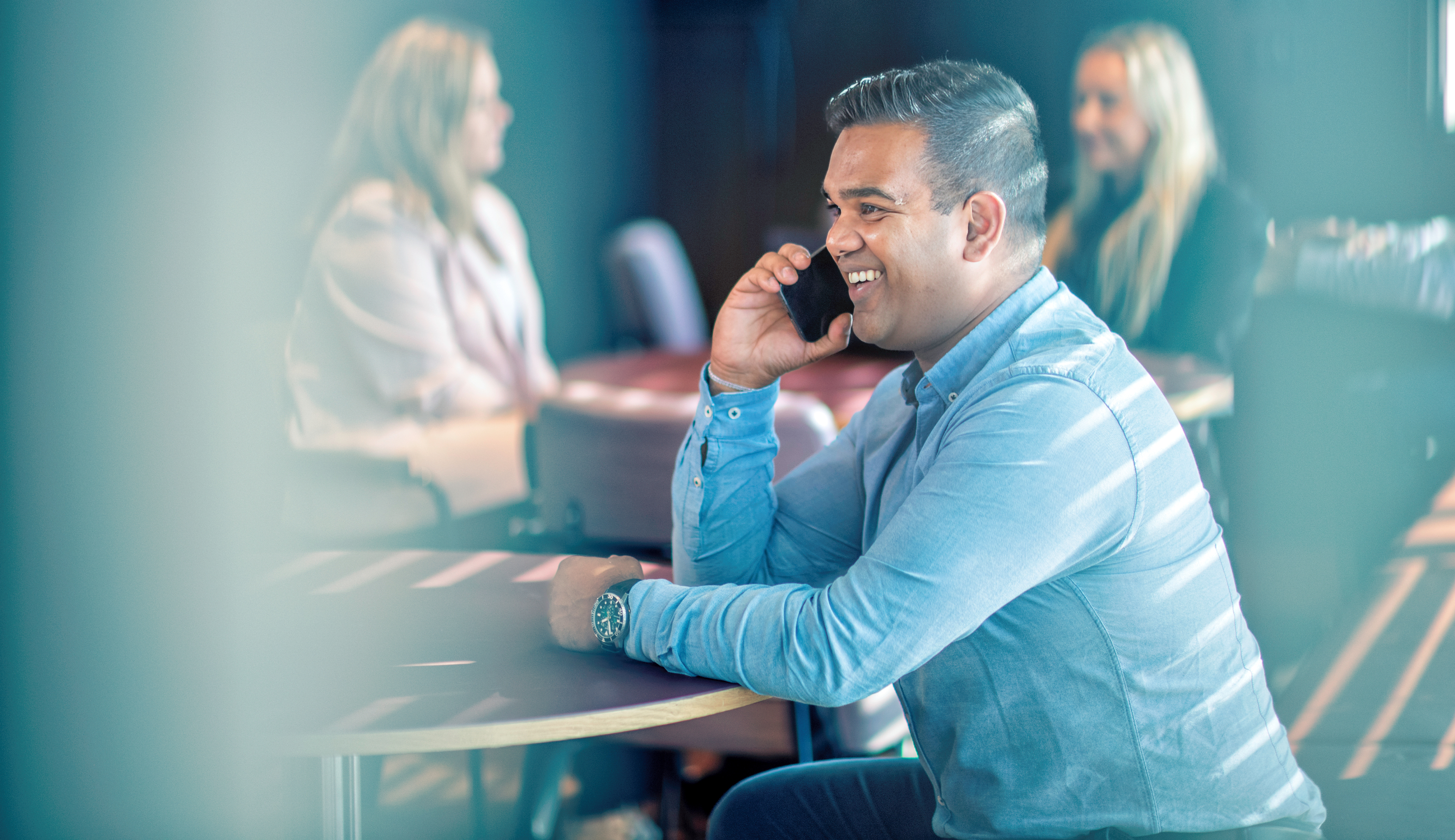 Smiling young man in blue shirt talking on the phone. People in the background.