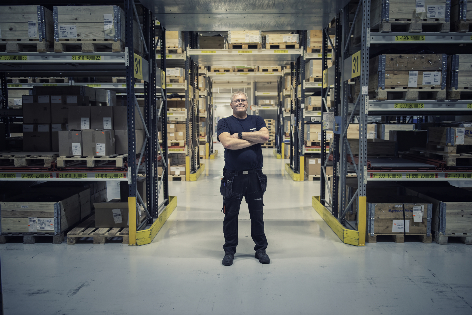 A male engineer stands in a warehouse surrounded by shelves of goods, smiling. The picture illustrates a typical workplace where occupational pensions can be an important part of the employees' future finances.