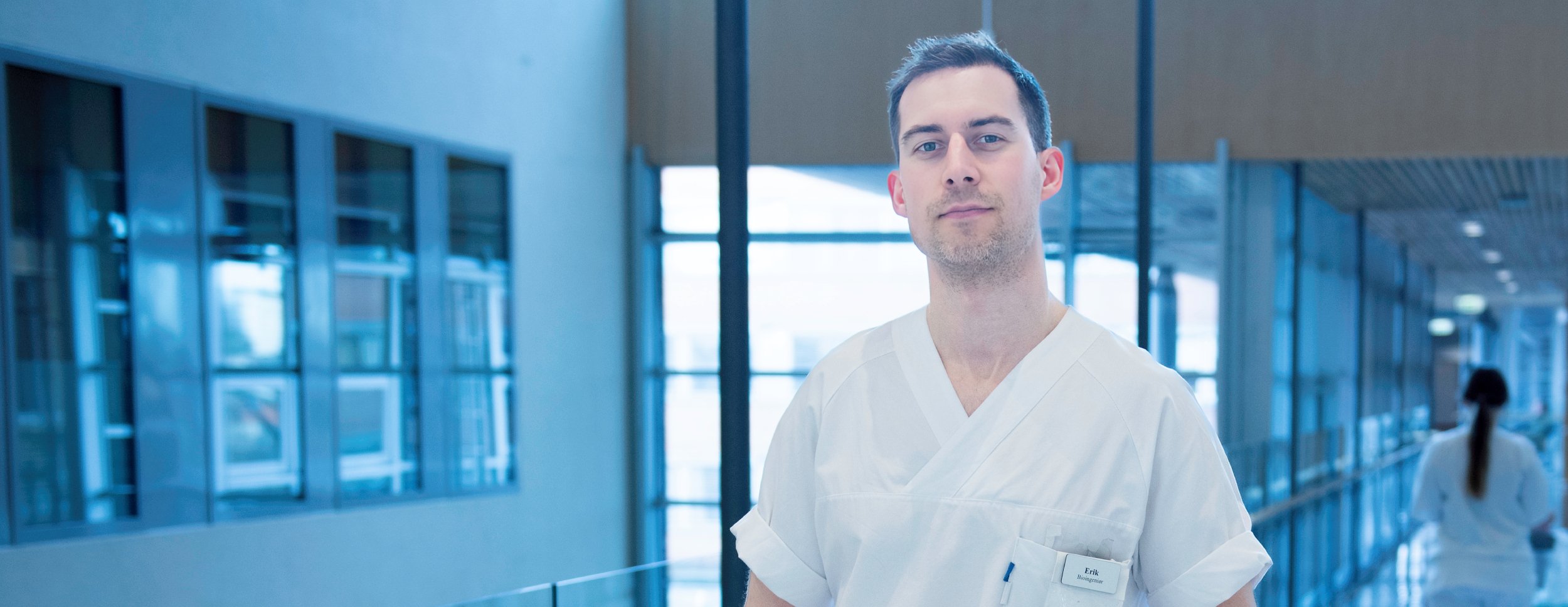 Male bioengineer in a white coat standing in a modern, bright hallway with large windows and glass walls. The person has a name tag on their chest. In the background, another person in a white uniform can be seen moving away from the camera.
