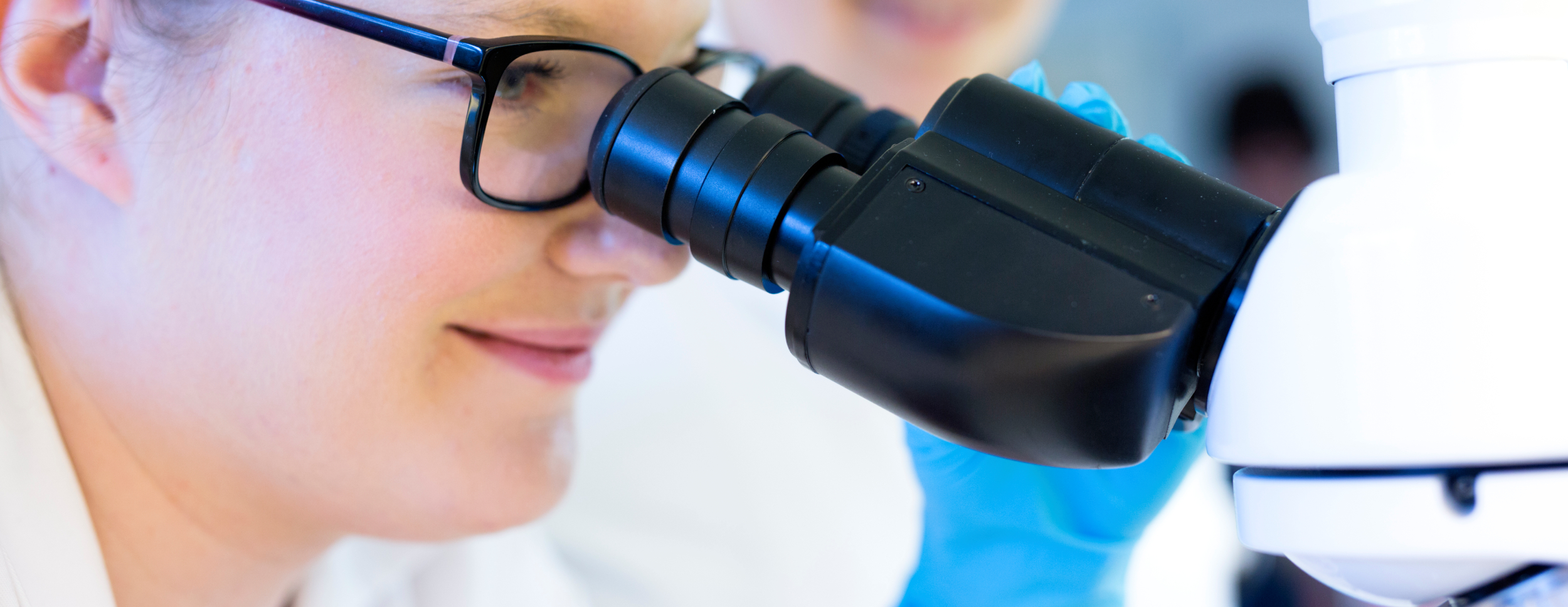 A person with glasses looks intently into a microscope. The image shows details of the face and the microscope.