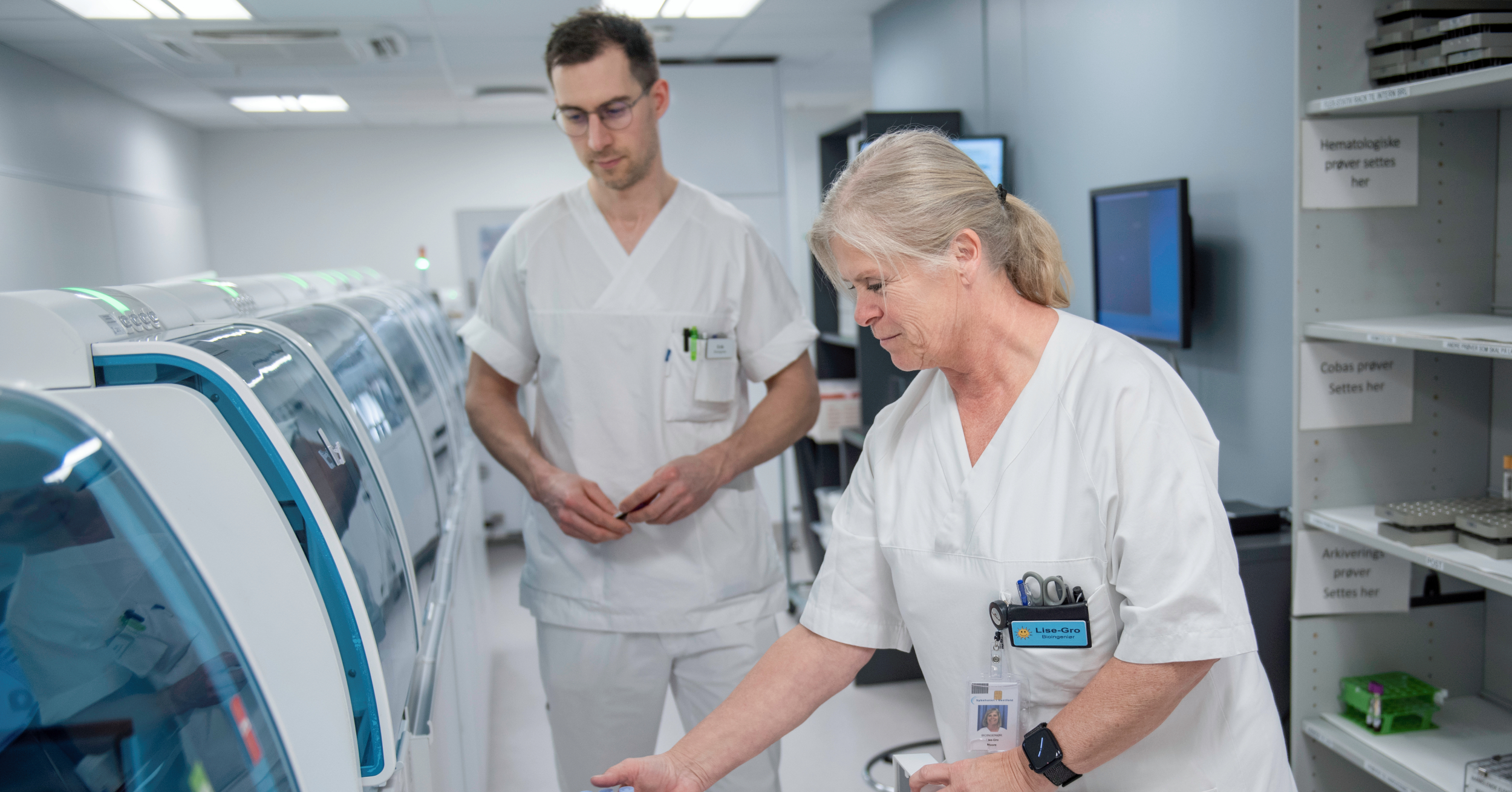 Two people in white lab clothes handle samples in a modern laboratory, at a large machine for medical or scientific analysis.