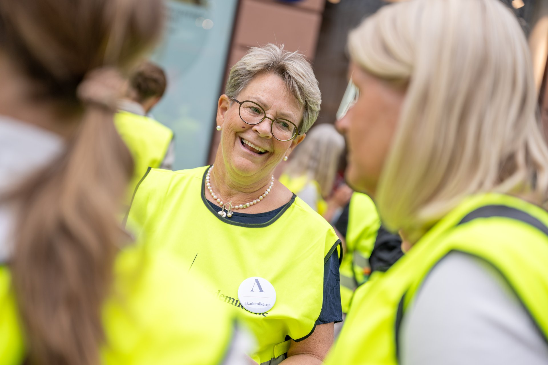An older member smiles as she participates in a strike, she is surrounded by other members in yellow vests and in good spirits.