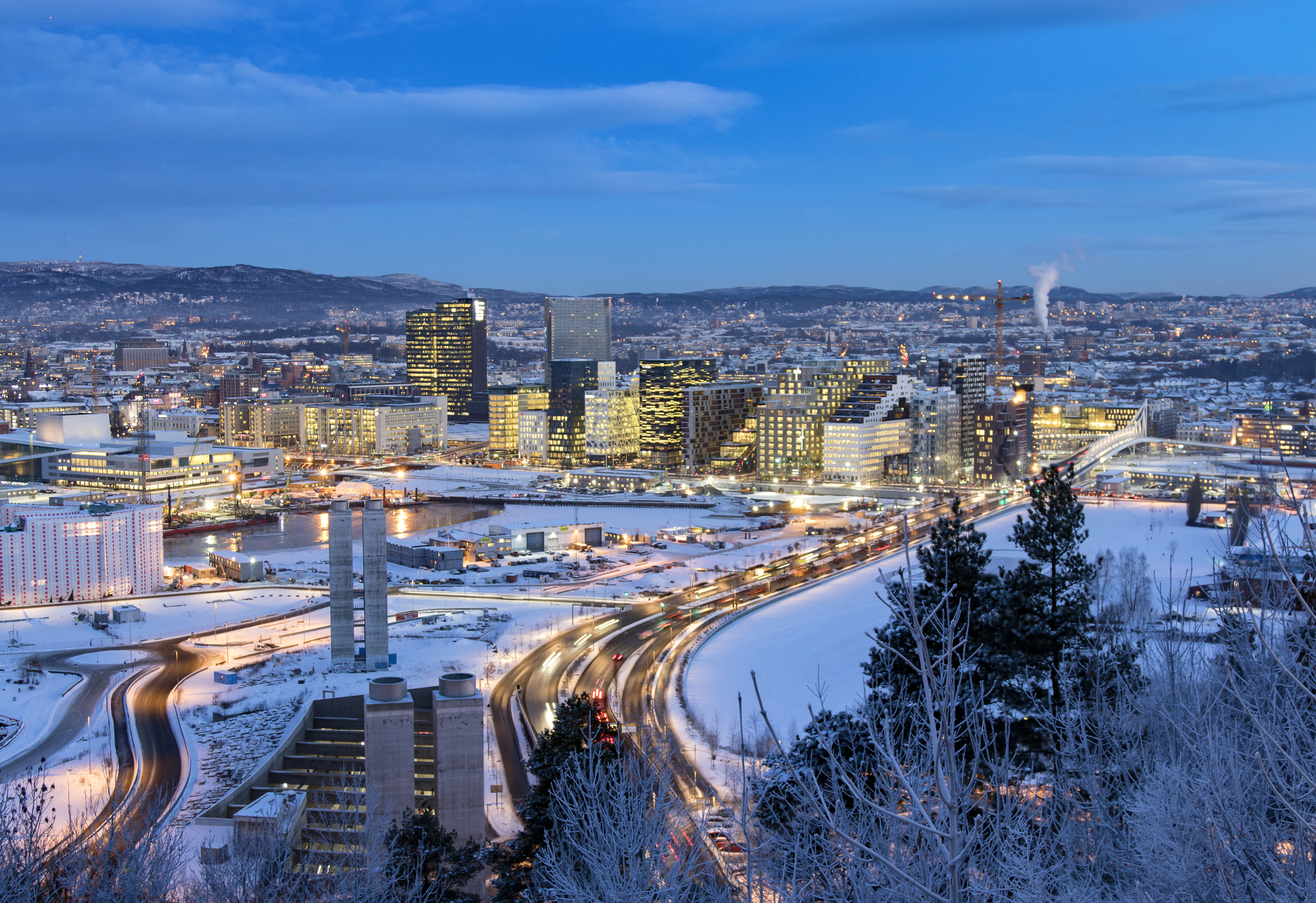 Utsikt over Oslo sentrum en vinterkveld, med snødekte gater, opplyste bygninger og blå himmel i bakgrunnen.