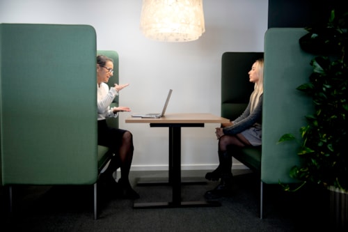 Two ladies are sitting in an open-plan office facing each other.
