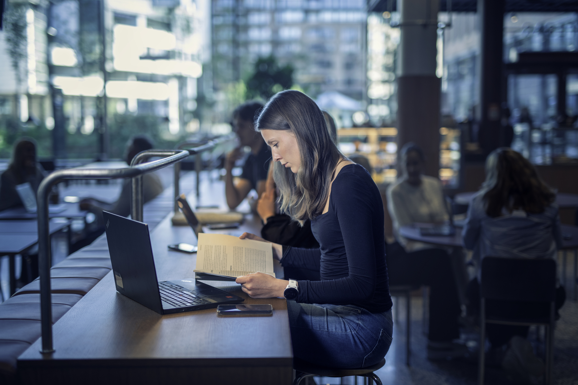 Ung kvinne studerer ved et bord i et moderne, lyst bibliotek, omgitt av bøker, laptop og andre studenter.