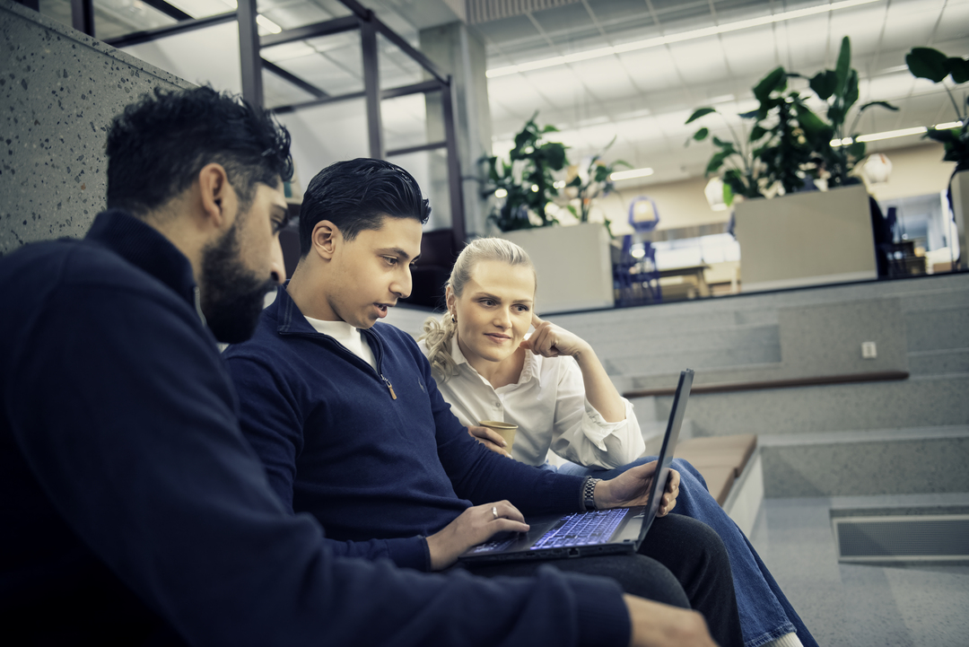Tre personer sitter sammen på en trapp innendørs, ser på en bærbar PC og smiler.