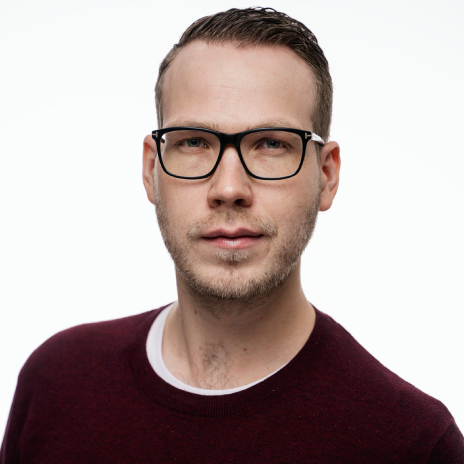 Man with glasses and a light beard, wearing a burgundy red sweater. Looking straight at the camera. The background is white.